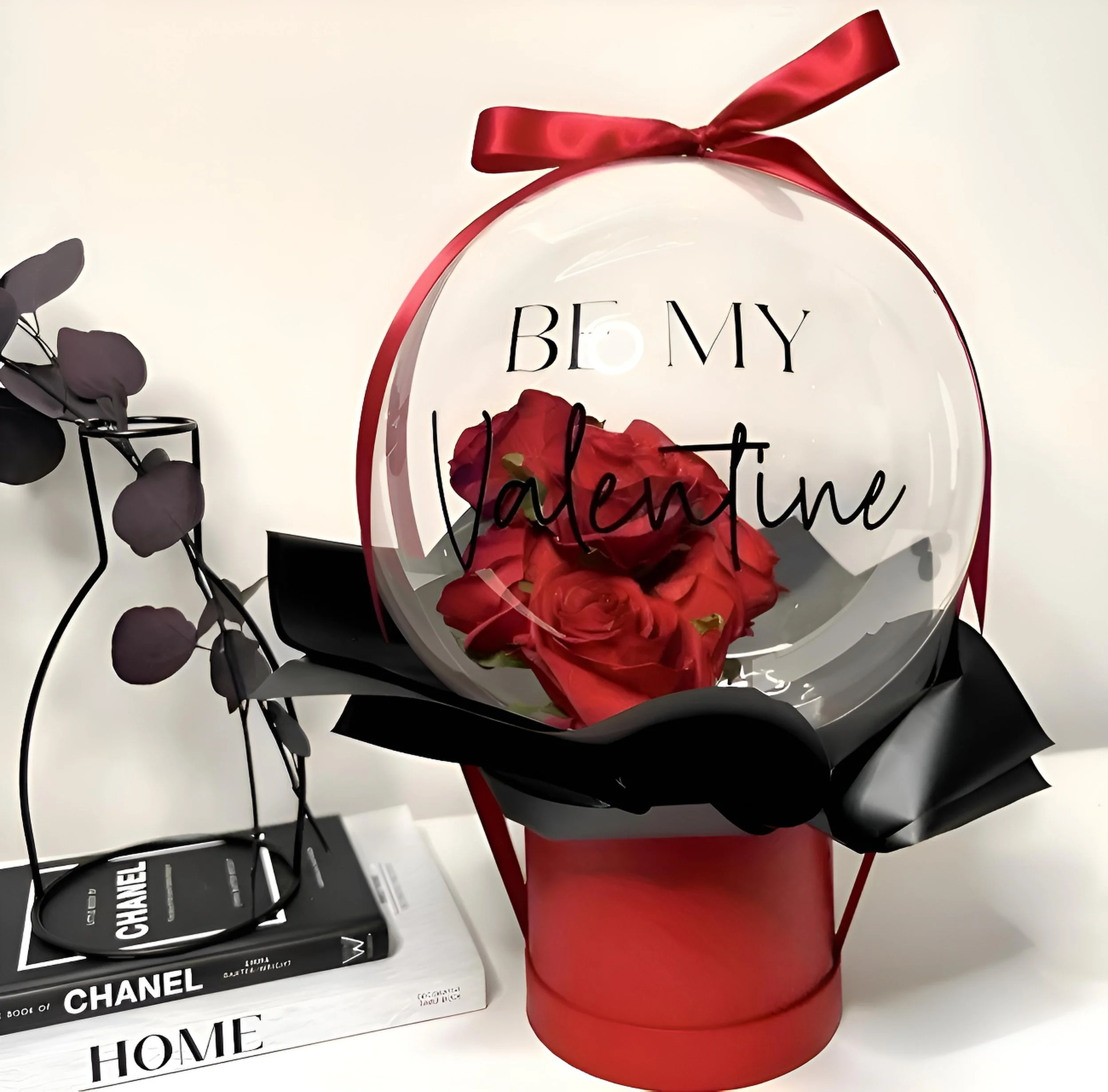 A Valentine's Day gift display with a bouquet of red roses inside a transparent ball with the words 'Be My Valentine' written on it, topped with a red ribbon. Next to it, a stack of fashion and home decor books with a black wire sculpture on top.