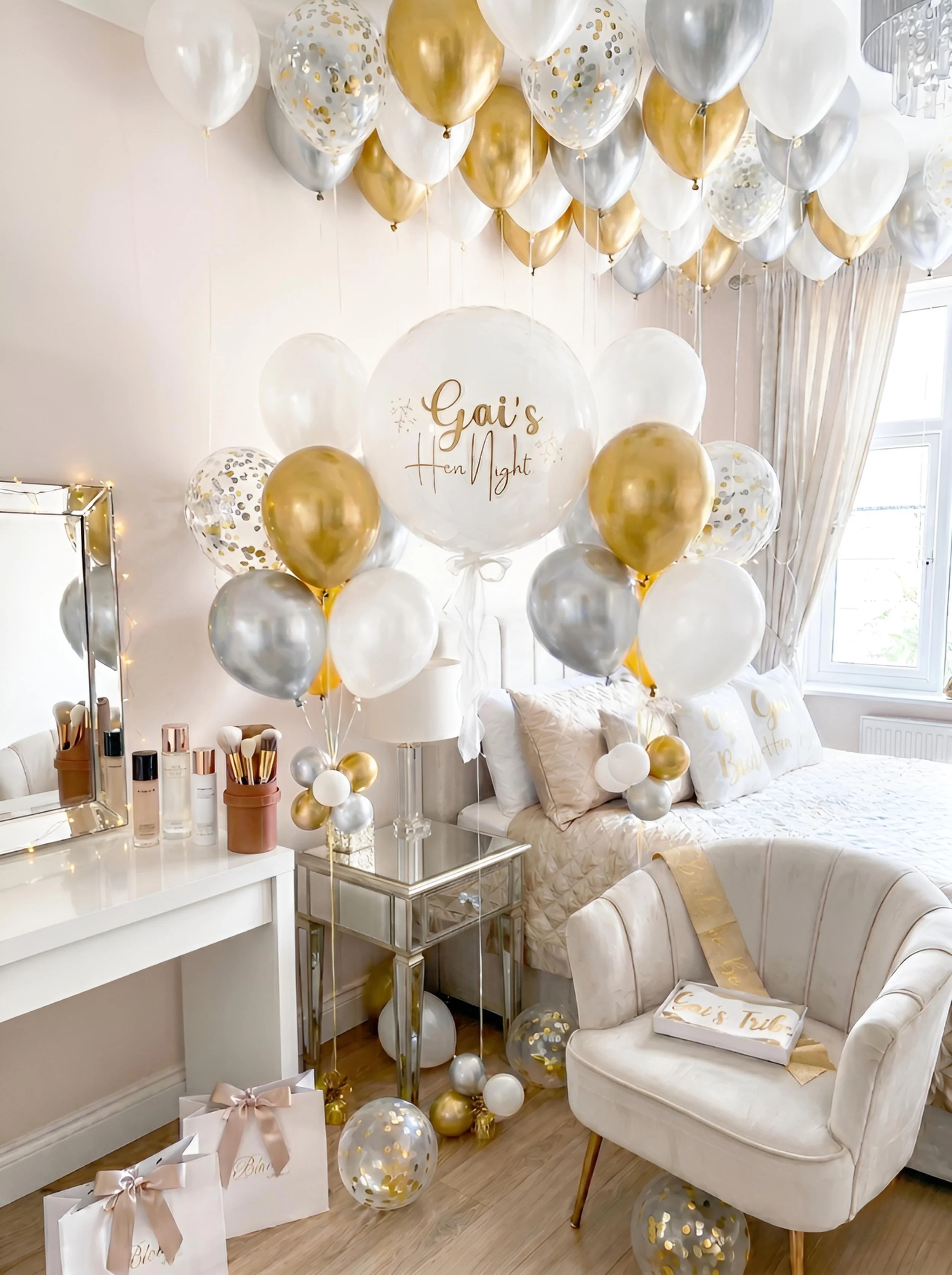 Room decorated with numerous white, gold, and silver balloons for a celebration. A large clear balloon in the center reads 'Gai's Hen Night' in gold lettering. There are gift bags with gold ribbons, a tray with makeup products, and a cozy armchair with pillows and a blanket in the background. Sunlight streams in through the window.