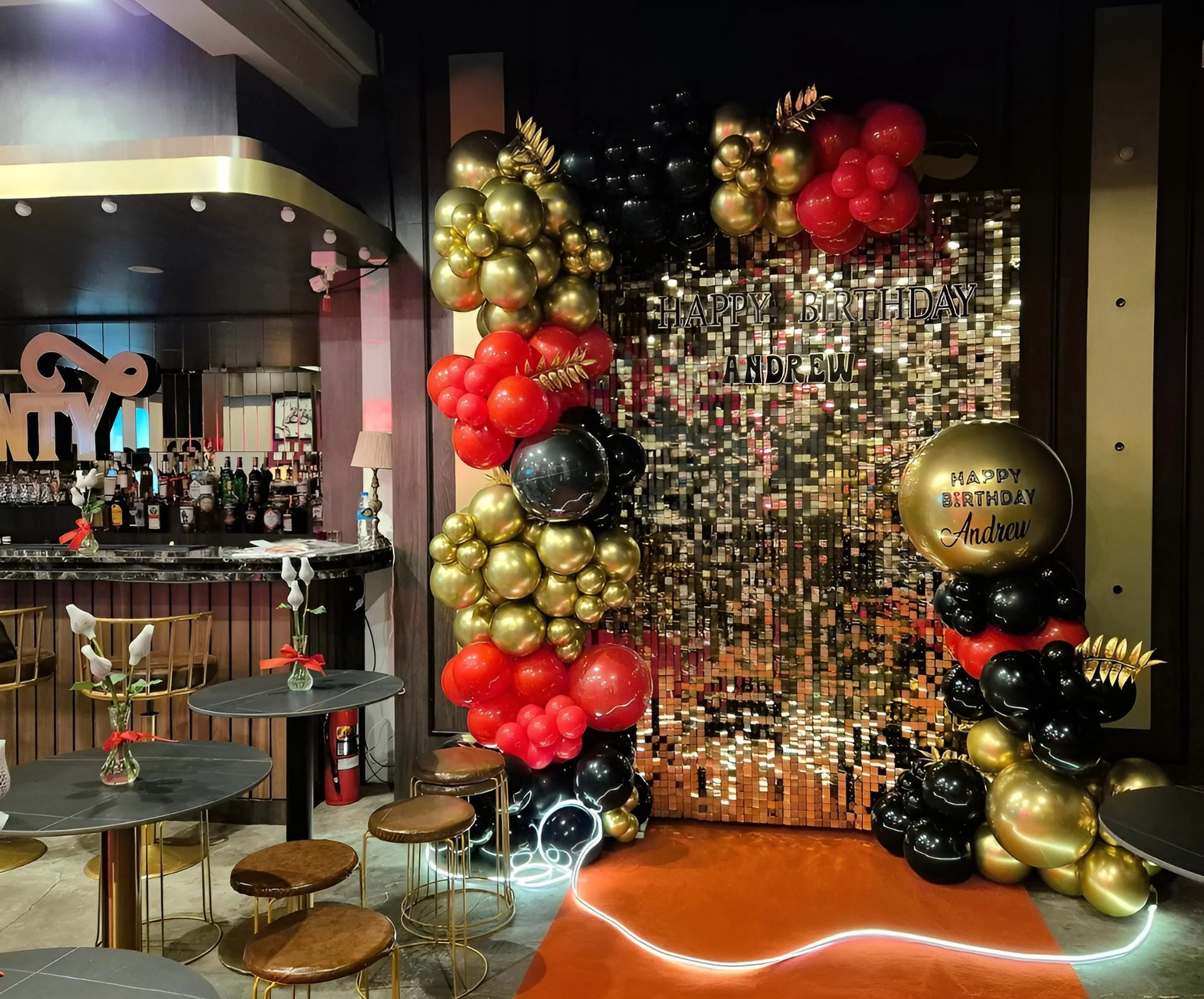 Decorative birthday celebration setup with a backdrop of gold and black sequined panels, balloons in red, black, and gold, and a sign that reads 'HAPPY BIRTHDAY ANDREW' in metallic letters, inside a restaurant or event space.