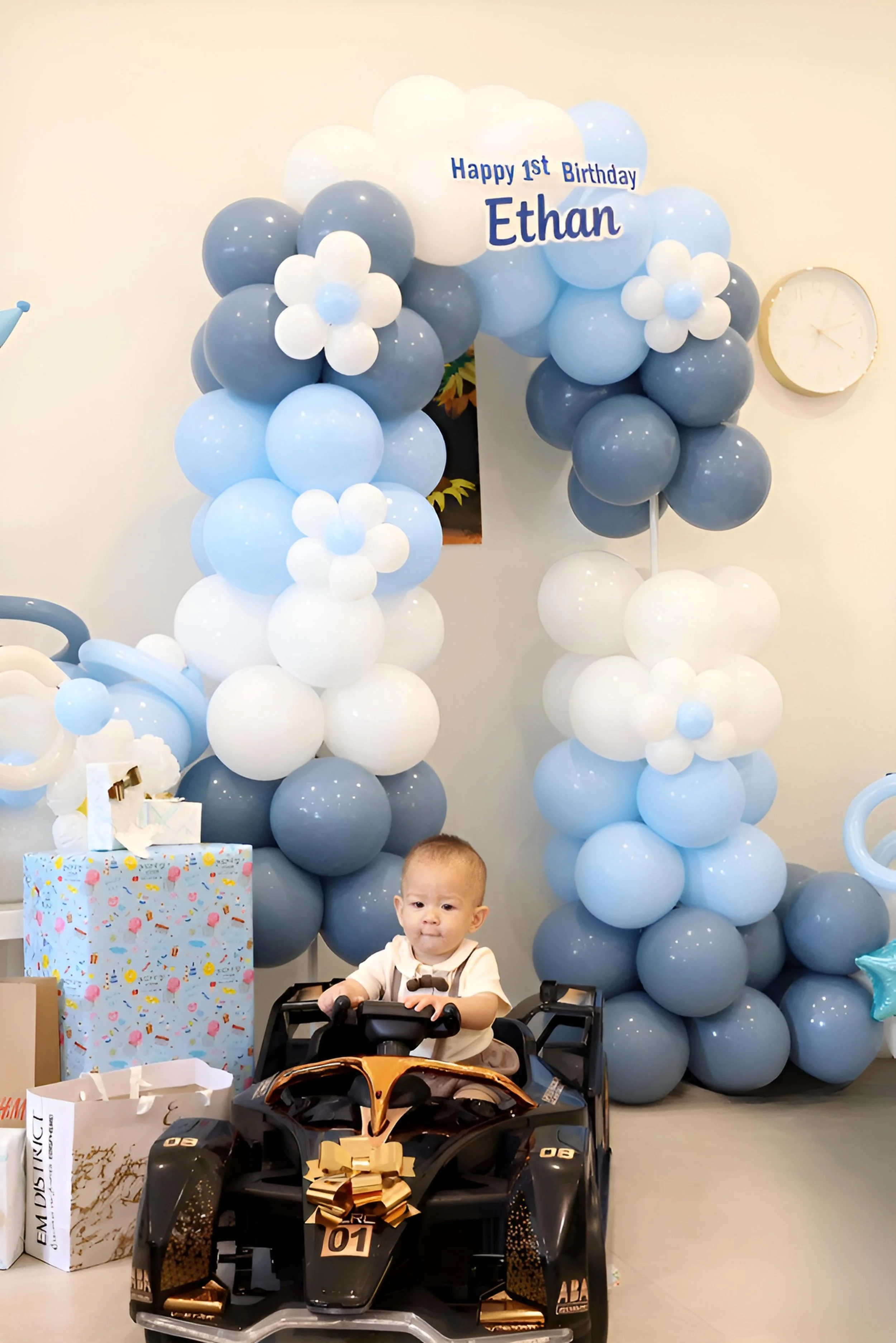 A young child sitting in a black and gold ride-on toy car, surrounded by birthday decorations including a balloon arch in shades of blue and white, birthday presents, and a birthday sign that says 'Happy 1st Birthday Ethan'.
