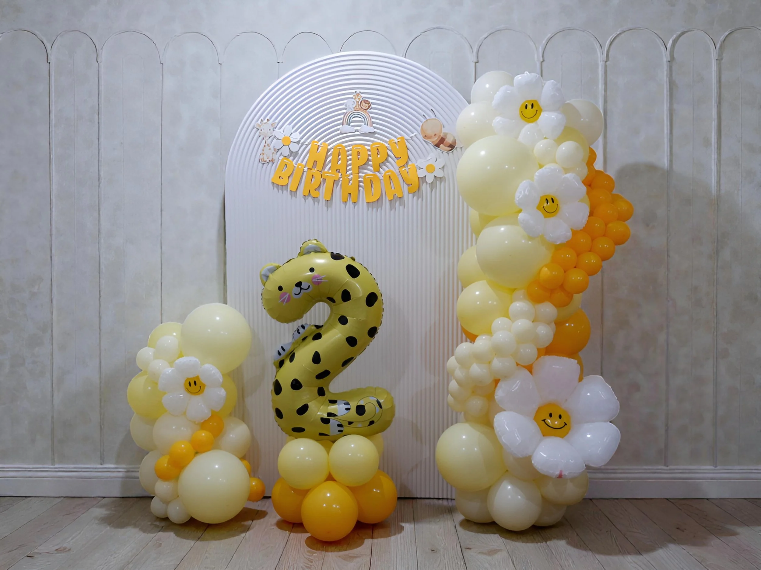 Birthday decoration with balloons, including a large number 2 balloon with a cheetah print, surrounded by white and yellow balloons, with some balloons shaped like flowers with smiley faces. There is a backdrop with a "Happy Birthday" sign and cute animal and rainbow illustrations.