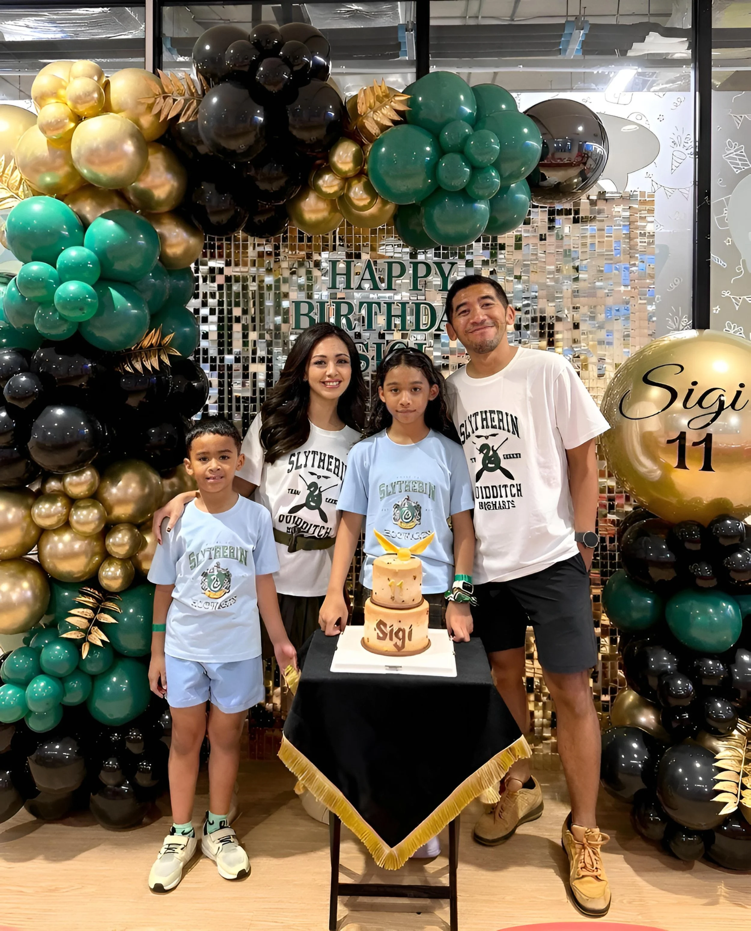 A birthday celebration with a woman, three children, and a man standing around a tall birthday cake with a gold pineapple on top. They are surrounded by balloon decorations in black, gold, green, and gold with the words 'Happy Birthday' in the background. The woman and the man are wearing white t-shirts with a green logo, and the children are wearing light-colored shirts.