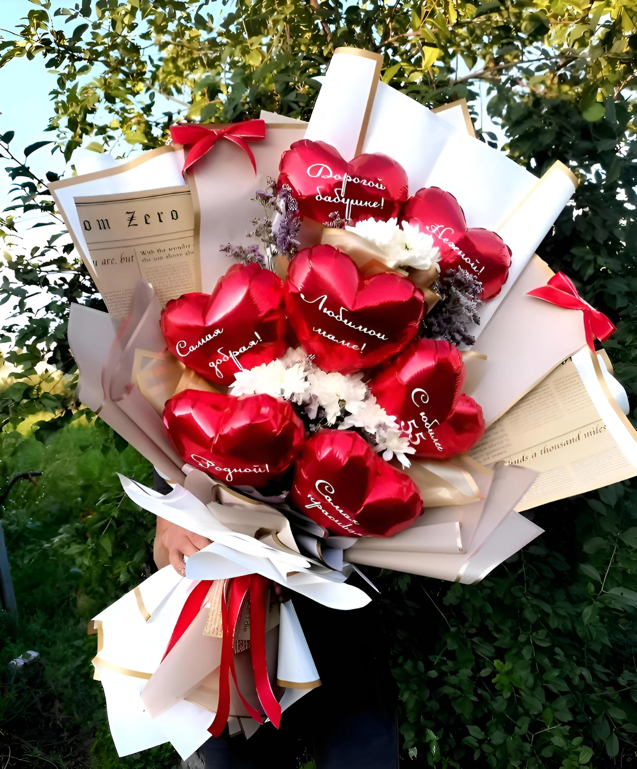 Person holding a bouquet of red heart-shaped balloons with white writing in Russian, surrounded by white and pink wrapping paper and flowers, outdoors with green foliage in the background.