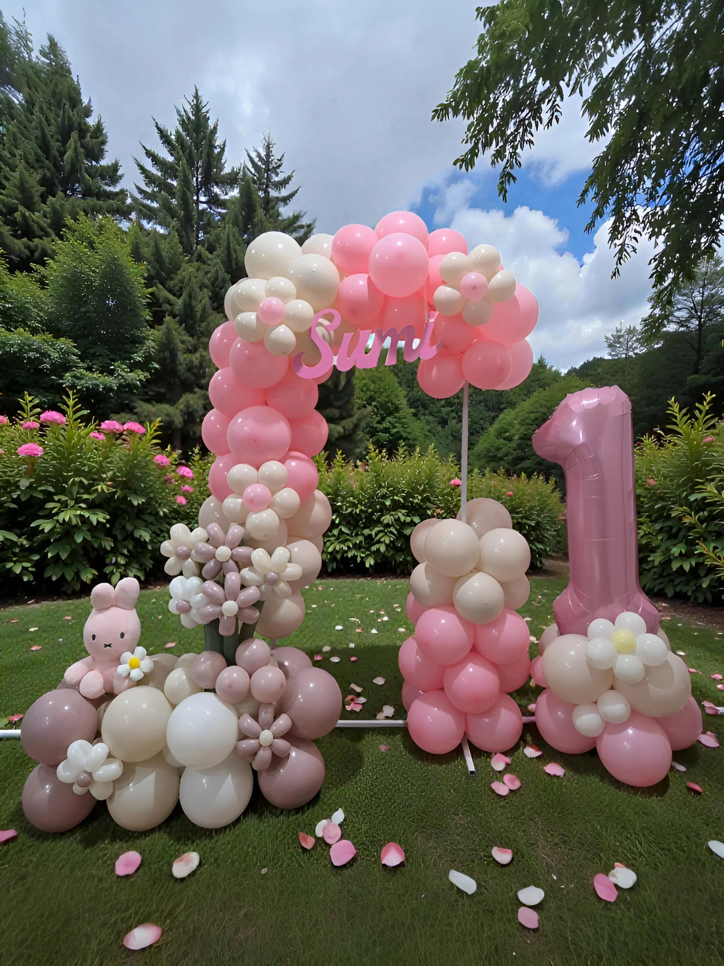 Decorative pink and white balloon arrangement with a pink number one balloon, pink and white flowers, a small plush bunny, and the word 'Sumi' in pink, set outdoors on a grassy area with trees and a partly cloudy sky in the background.