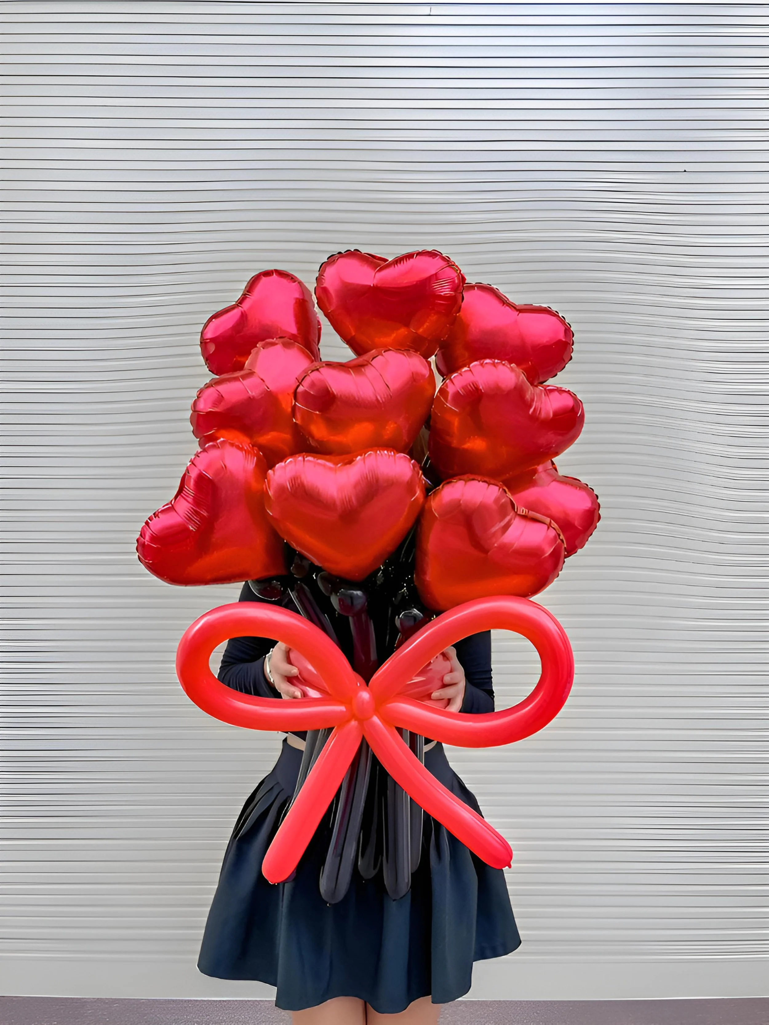 Person holding a large bouquet of red heart-shaped balloons with a red balloon bow at the base, against a gray horizontal line background.