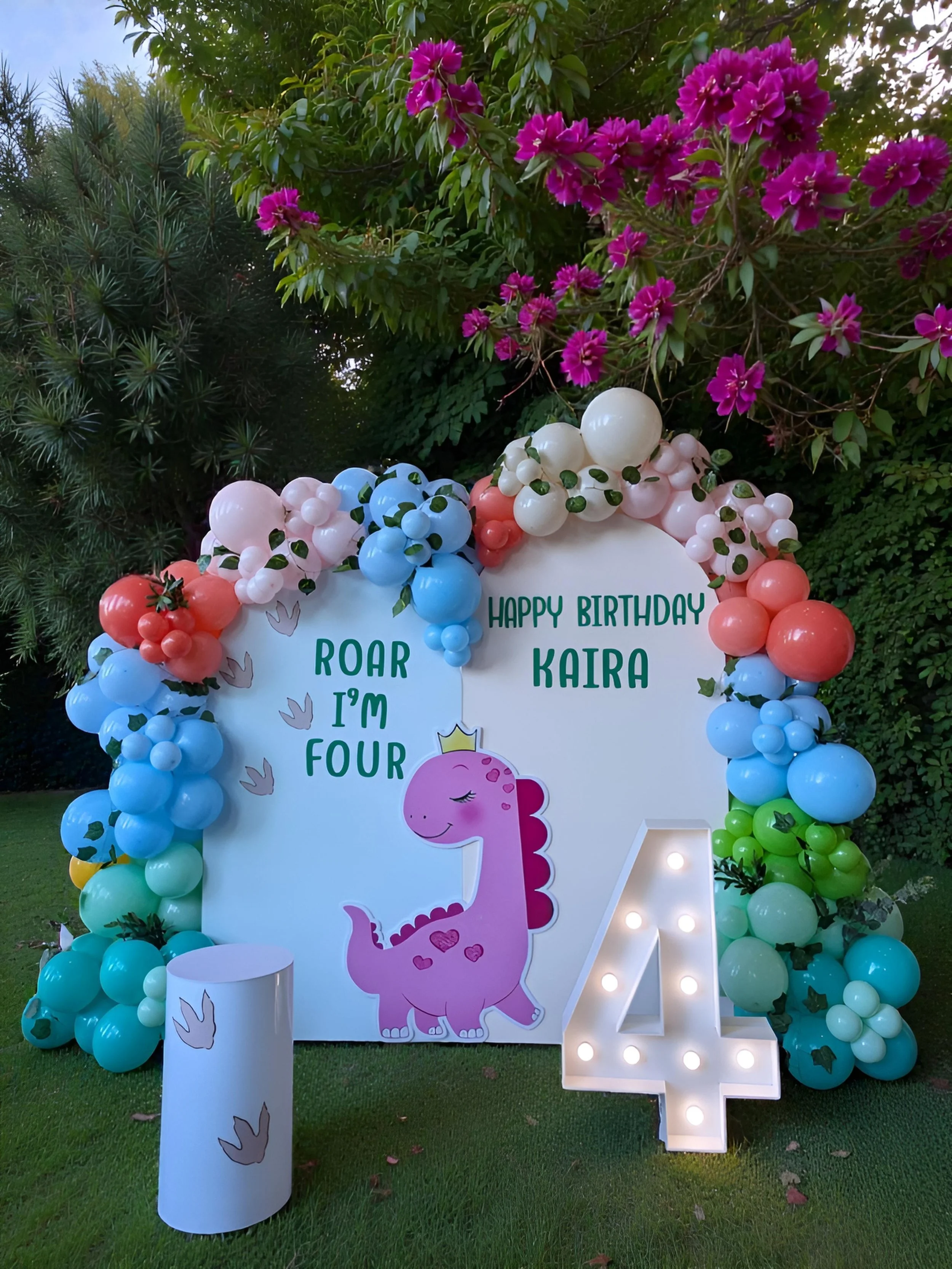Decorative birthday setup with balloon arch, a large illuminated number 4, and a white backdrop with a pink dinosaur cartoon. The backdrop has birthday wishes for Kaira, who is turning four, with the words 'Roar I'm Four' and 'Happy Birthday Kaira.' The balloon arch consists of pink, white, blue, and green balloons, with greenery accents. A cylindrical white stool with bird illustrations is in front of the backdrop, set on a grassy area with trees and pink flowers in the background.