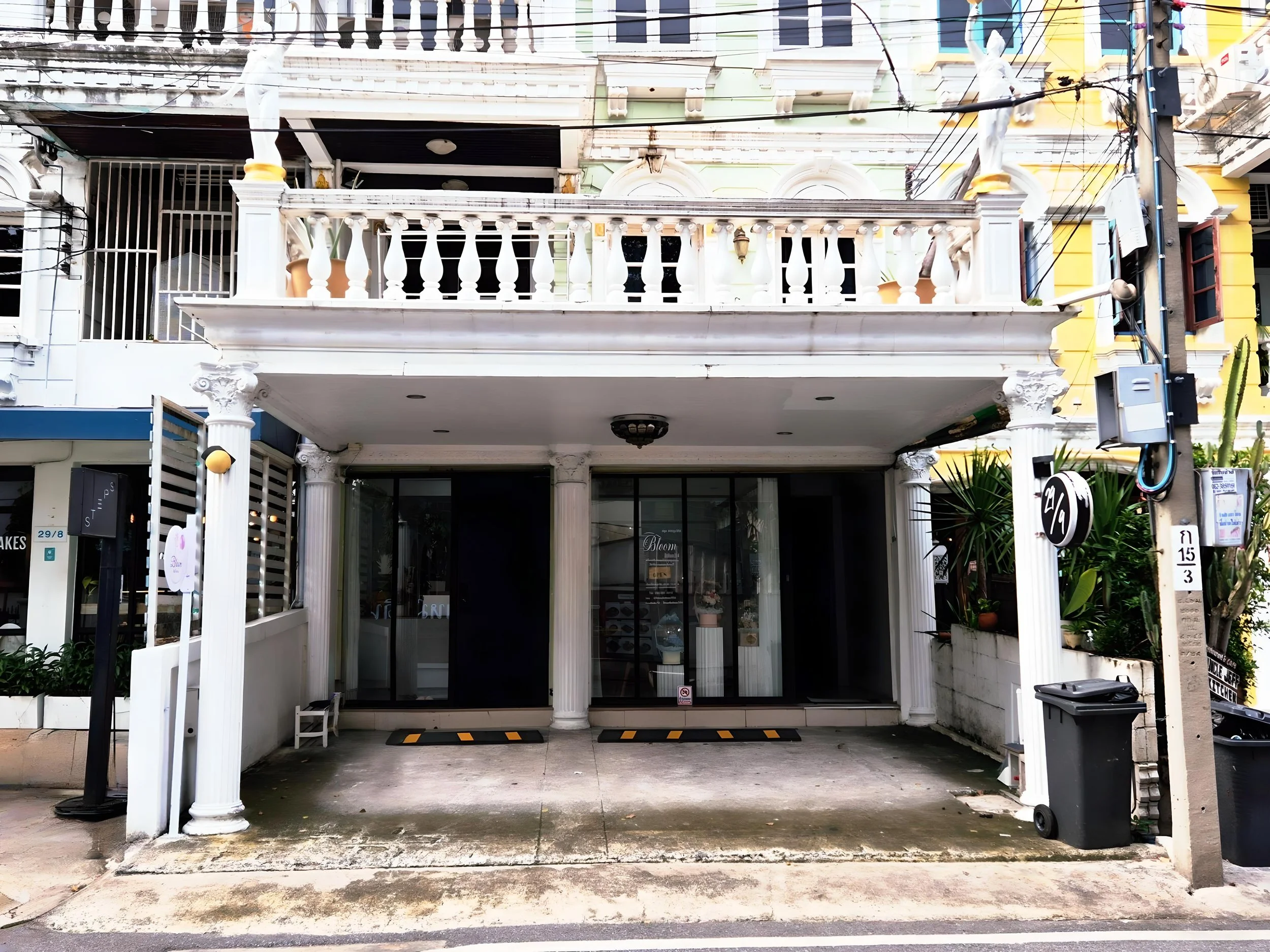 Front view of a building with classical white columns and ornate balcony railings, featuring a garage-style entrance and surrounding plants.