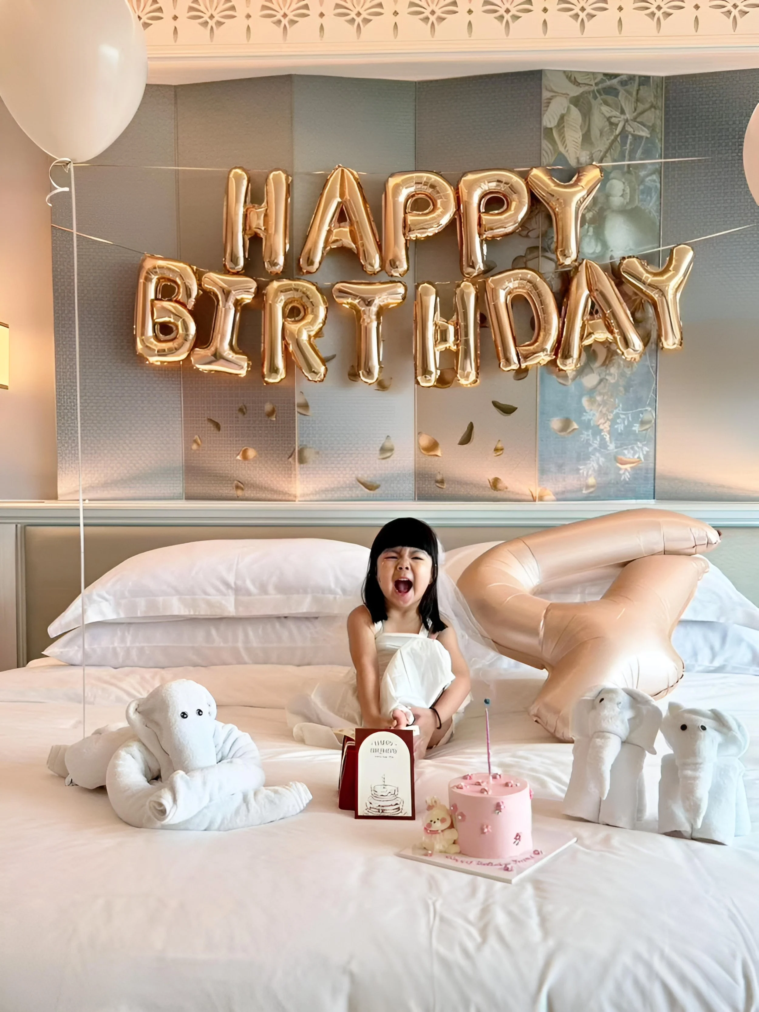 A young girl celebrating her birthday with a 'Happy Birthday' balloon banner, teddy bears, and a pink cake with a single candle, in a decorated room with white bedding and soft plush toys.