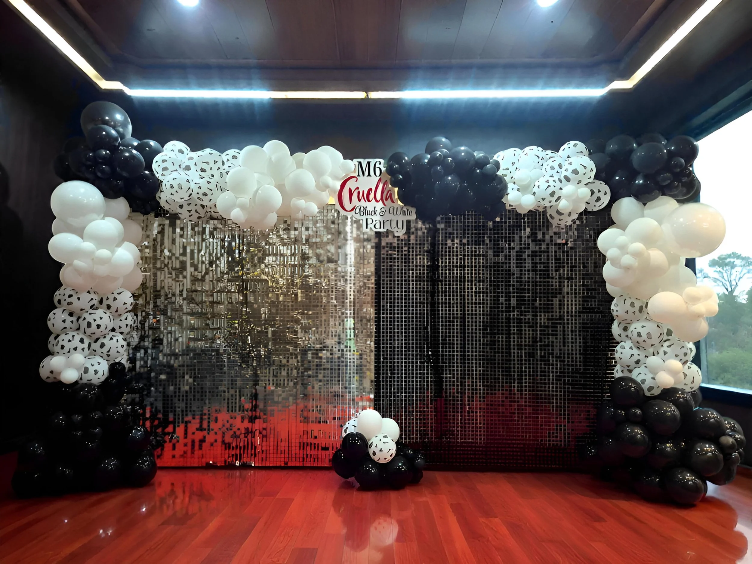 Balloon arch with black, white, and black-and-white patterned balloons, decorated with a sparkling reflective backdrop at a party, with a sign that reads 'Cruella' and 'Club & White Party'.