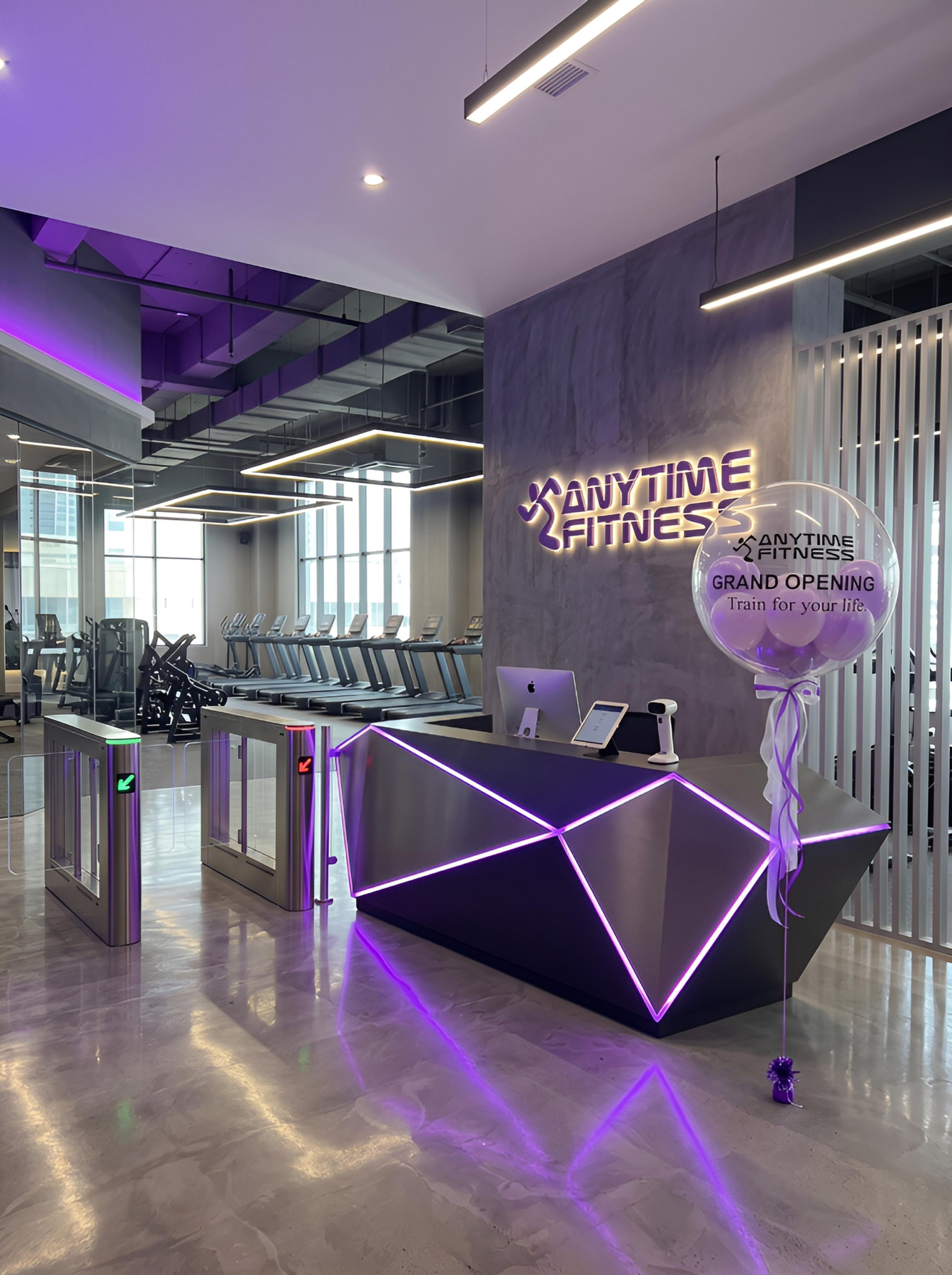 Interior of a modern fitness center with a reception desk, illuminated with purple LED lighting. Treadmills are lined up along large windows in the background. There's a helium balloon with a grand opening message and a small gift box. The ceiling has geometric light fixtures, and the space features marble walls and flooring.