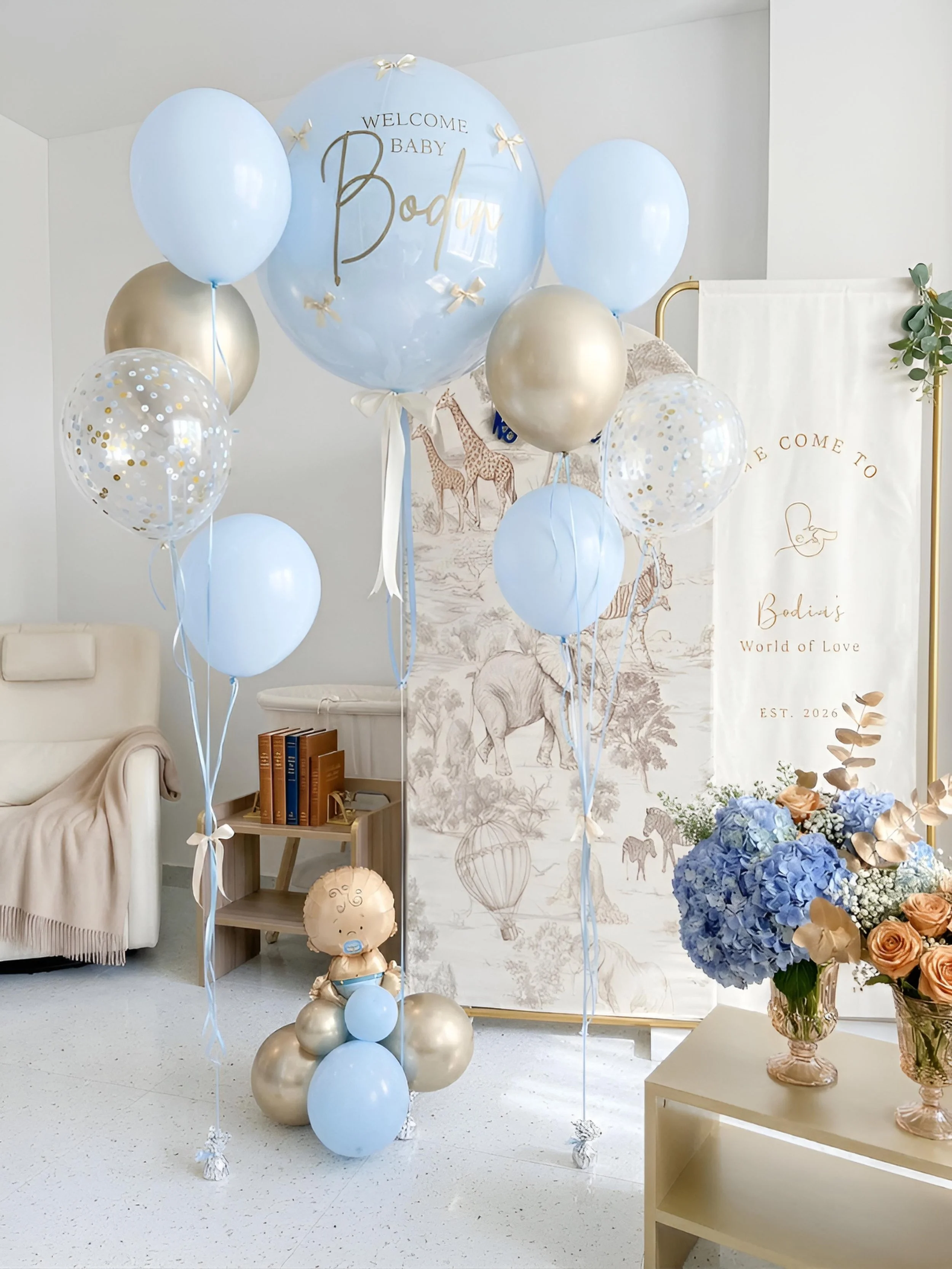 Baby shower decor with a large blue transparent balloon that says "Welcome Baby Bodhi" and smaller balloons in blue, gold, and clear with confetti. There are floral arrangements with blue and peach flowers in a pink vase, a white banner with gold text and animal illustrations, and a wall mural featuring giraffes and elephants.