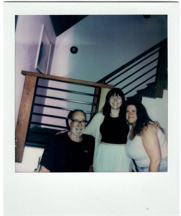 Three people smiling and posing together indoors in front of a staircase. The group consists of an older man with glasses and a beard, a woman with black hair and glasses, and a woman with long curly dark hair. The setting appears to be a home enviro