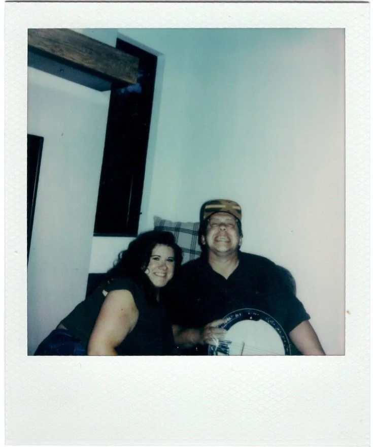 Two people, a woman with dark curly hair and a man wearing a cap, sitting close together indoors. The man is holding a musical instrument and smiling broadly, while the woman is also smiling.