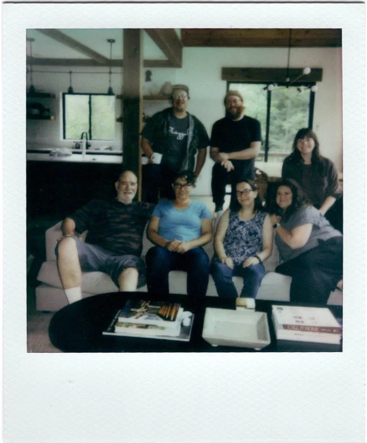 A group of six people gathered in a living room, with three seated on a sofa and three standing behind them, smiling for the camera. There are windows in the background showing greenery outside, and a black coffee table in front of the sofa with maga