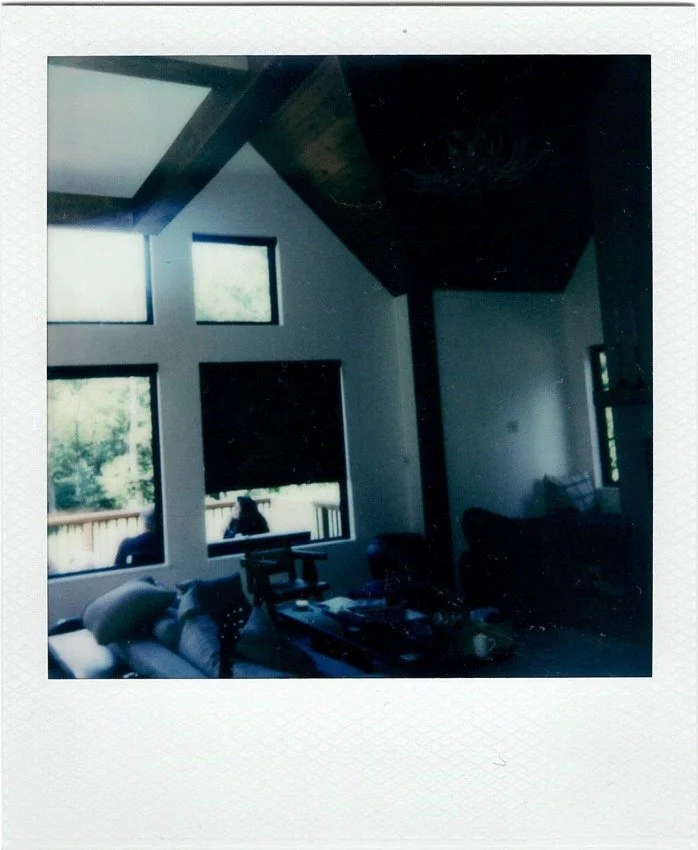 Interior of a living room with large windows, a sectional couch with pillows, a coffee table with various items, and a high ceiling with exposed beams.