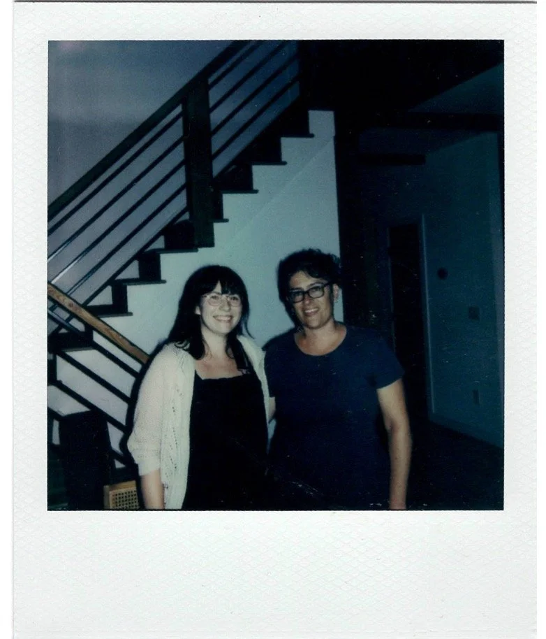 Two women with dark hair and glasses standing together indoors near a staircase, smiling at the camera.