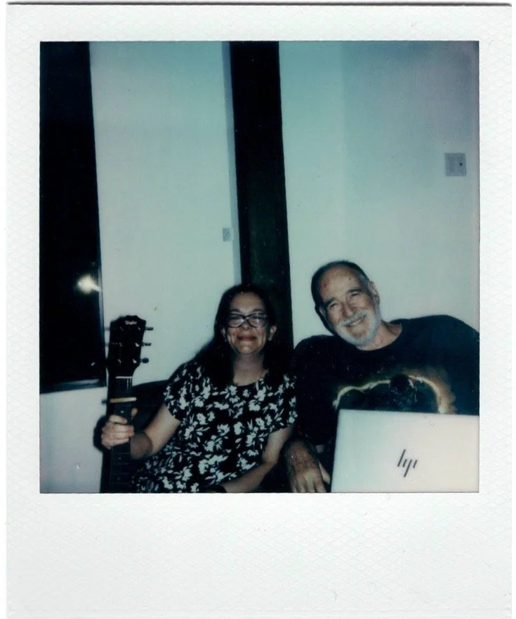 Two people sitting together, smiling, with the woman holding a guitar, and a laptop in front of the man, in a room with white walls.