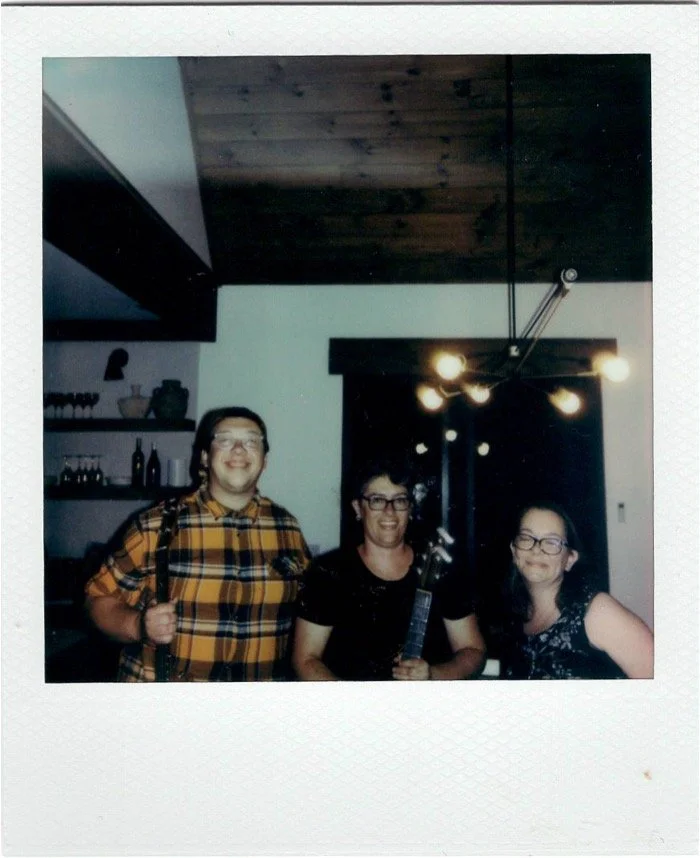 Three women standing inside a house, smiling at the camera; one woman has a backpack, and the woman in the middle is holding a bottle.
