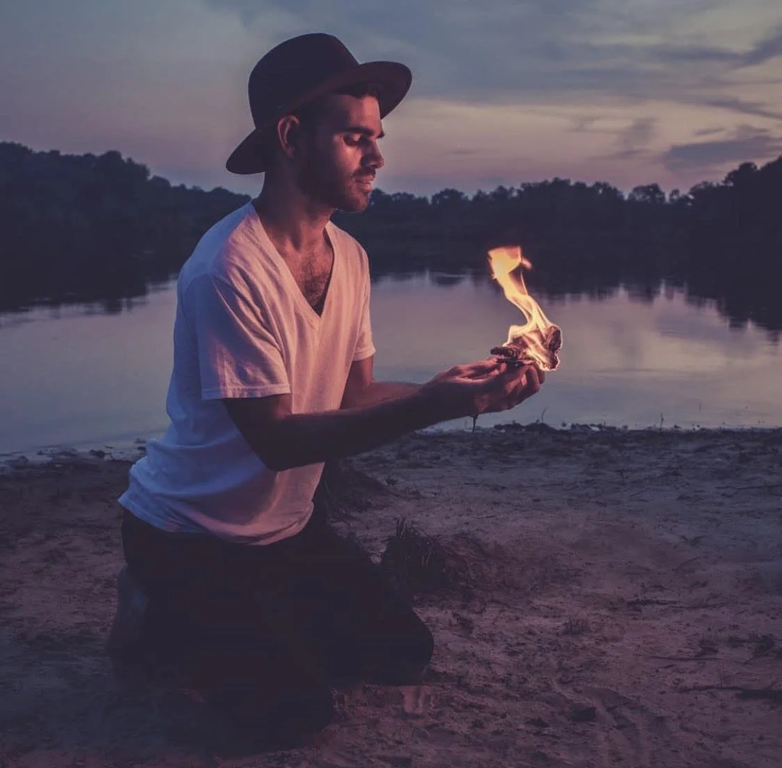 Man in white t-shirt and hat kneeling by a lake holding a small fire at dusk.