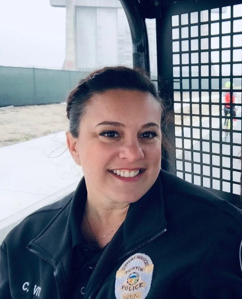 A smiling female police officer with dark hair, wearing a black uniform with a badge and the words 'Austin Police' on her shoulder, sitting in the back of a police vehicle.