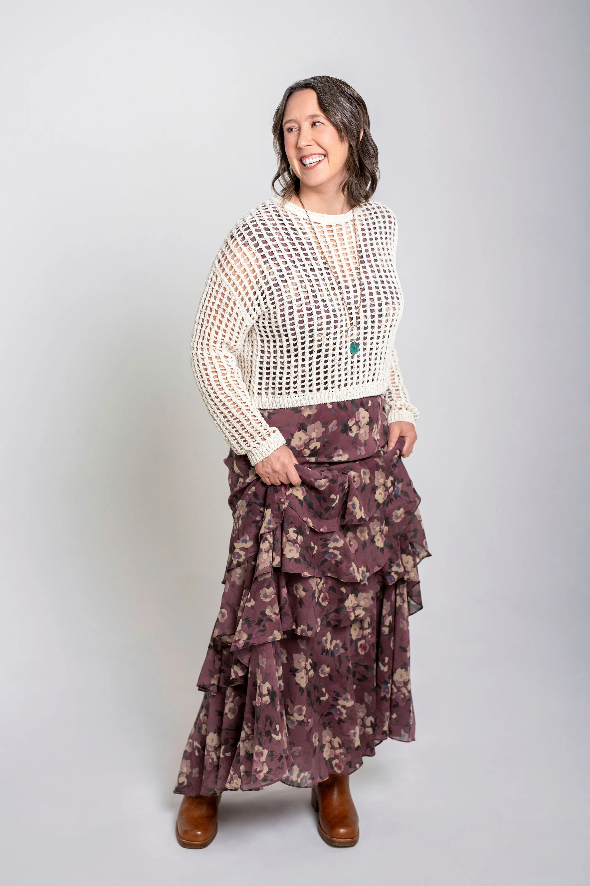 Jolee wearing a long purple skirt, brown boots, and an open weave cream sweater. She is laughing and looking over her shoulder.