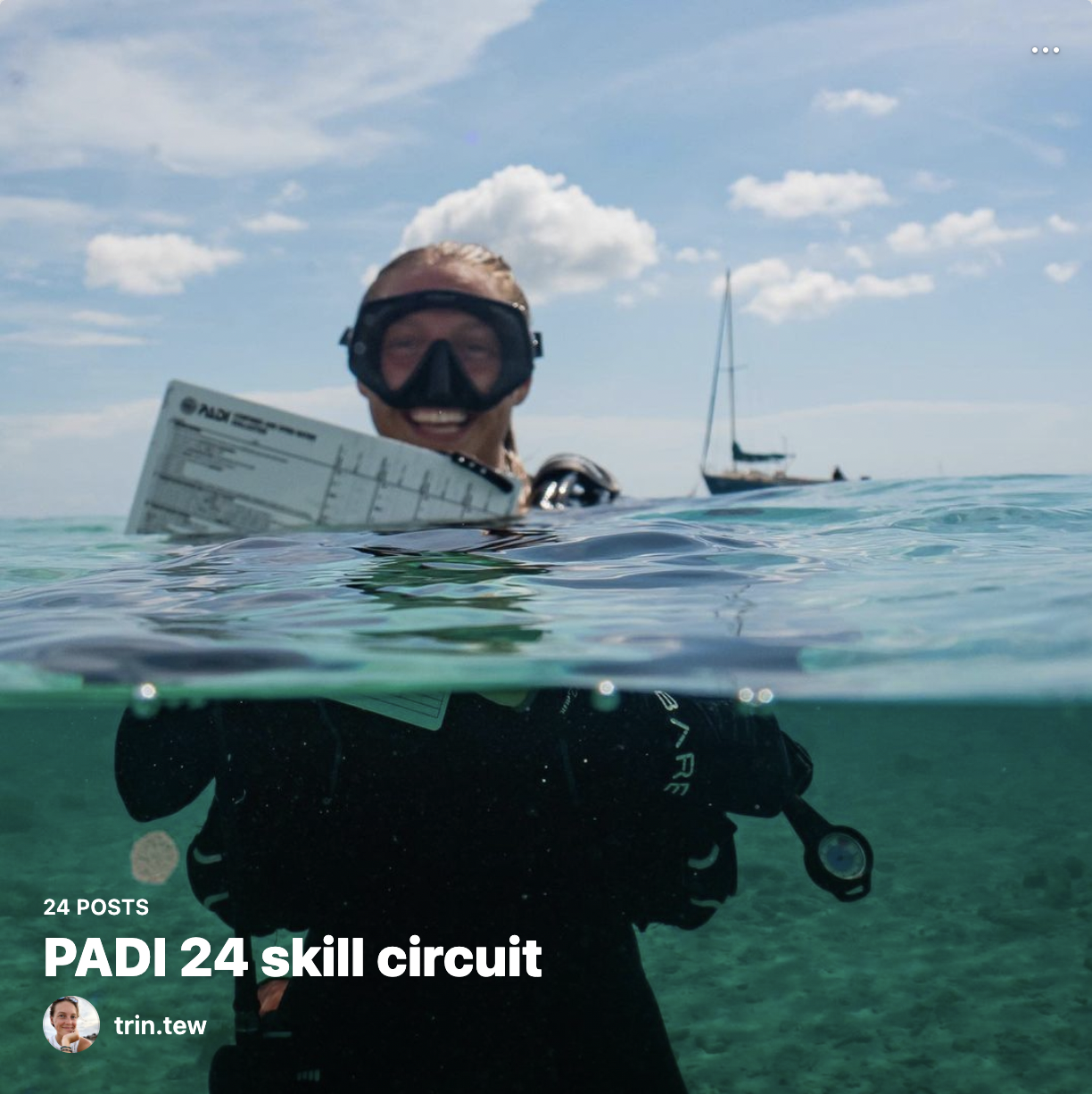 A person in a wetsuit with goggles swimming in the ocean, holding a PADI certification card, with a sailboat in the background under a partly cloudy sky.