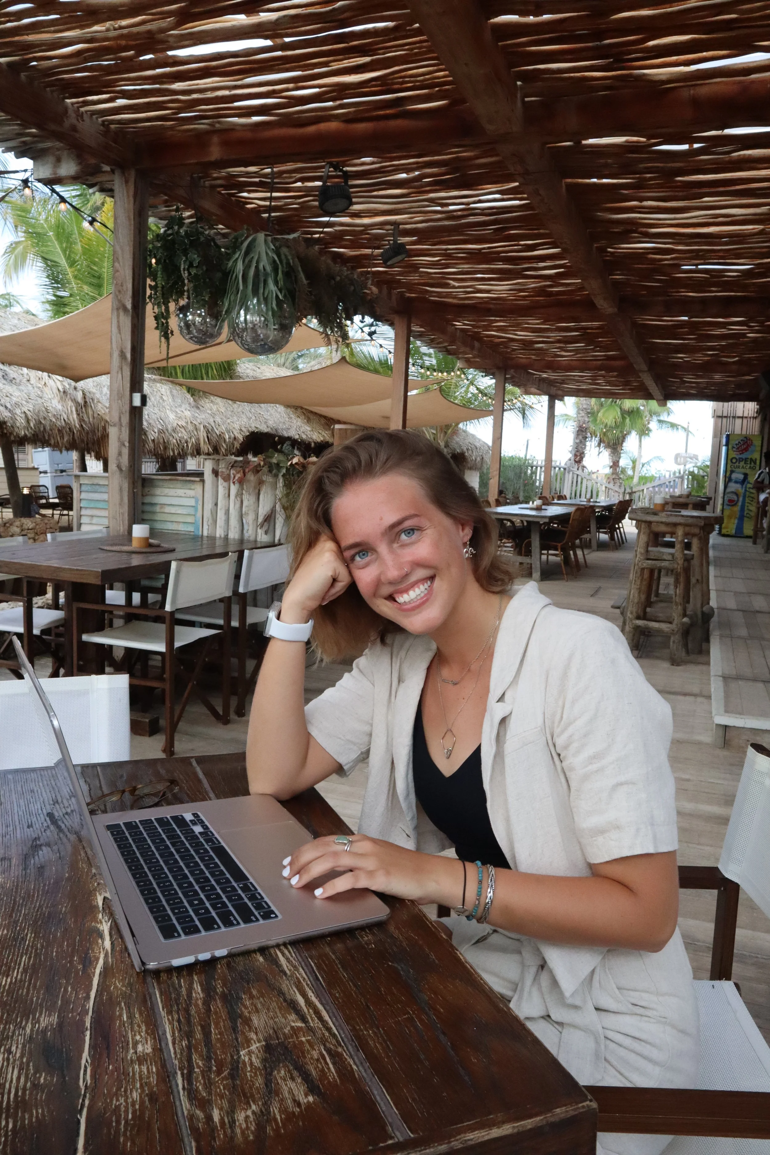 A young woman with blonde hair and blue eyes smiling while sitting at a wooden table with a laptop, in an outdoor tropical restaurant or bar setting with thatched roofs, palm trees, and string lights.
