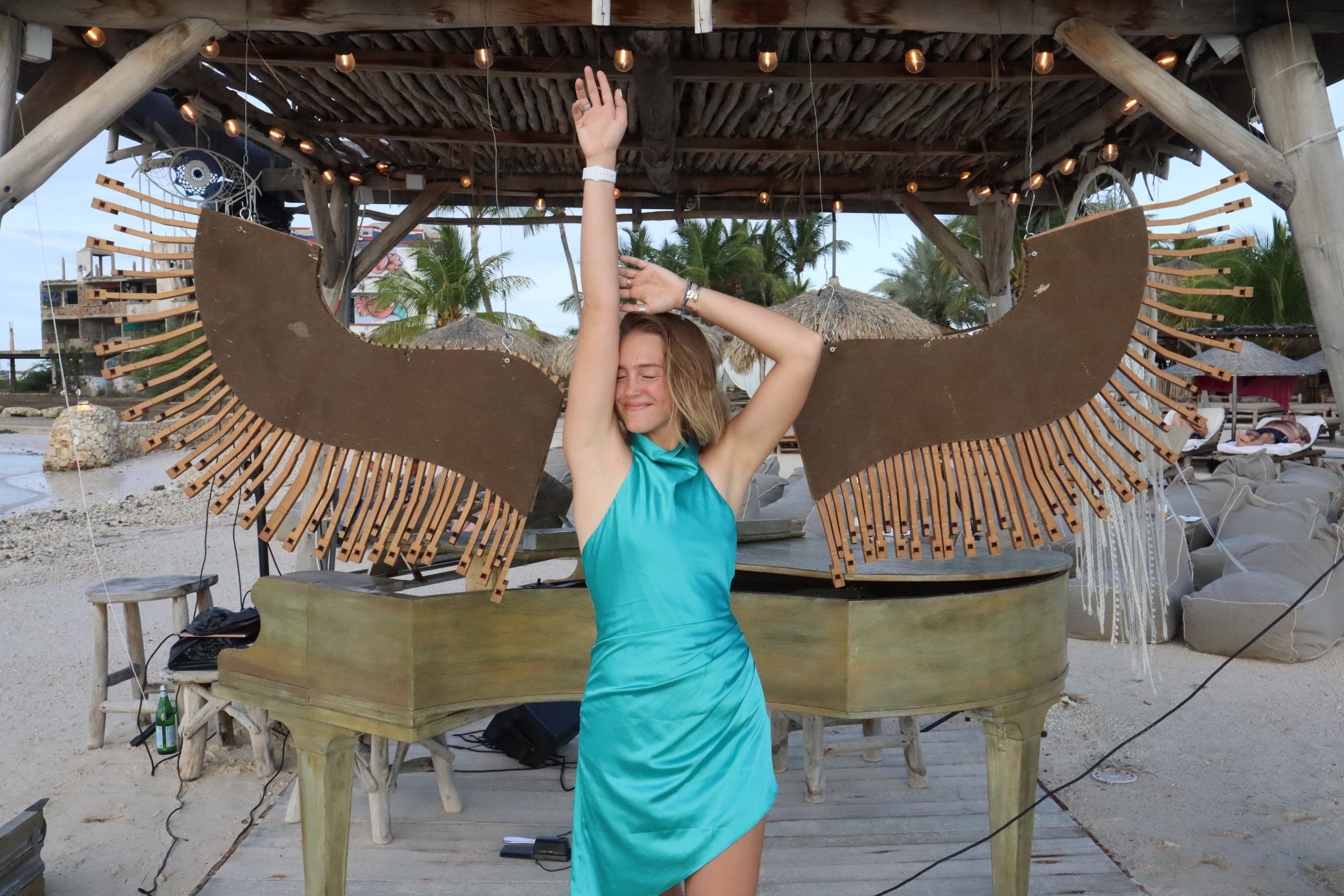 A woman in a turquoise dress dancing with an angel wings costume at an outdoor beach venue with palm trees and thatched umbrellas in the background.