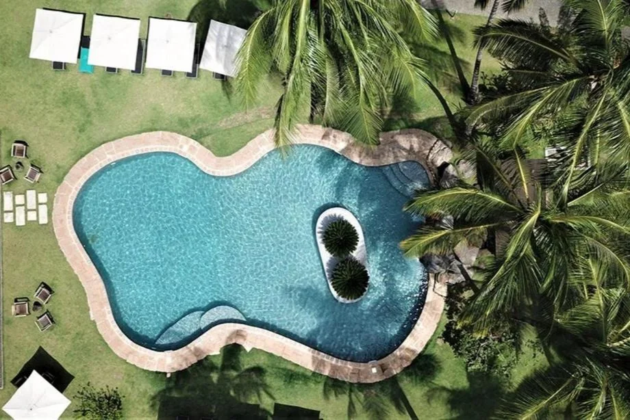 Vue aérienne d'une piscine en forme de cœur entourée de palmiers et de chaises longues dans un espace tropical.