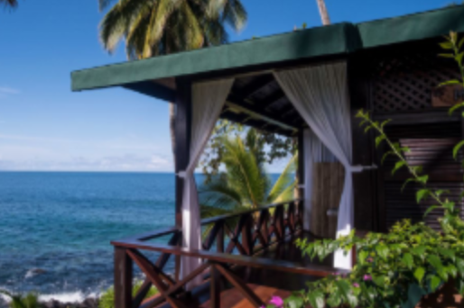 Terrasse en bois avec rideaux blancs, vue sur la mer et palmiers sous un ciel bleu