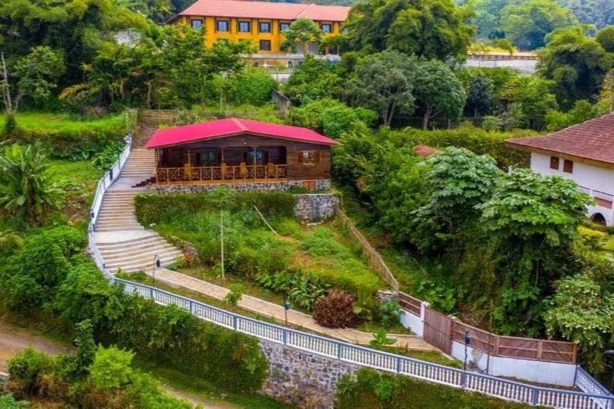 Une maison en bois avec un toit rouge, située sur une colline verdoyante entourée d'arbres, avec un escalier en pente et une rampe blanche menant à la maison.