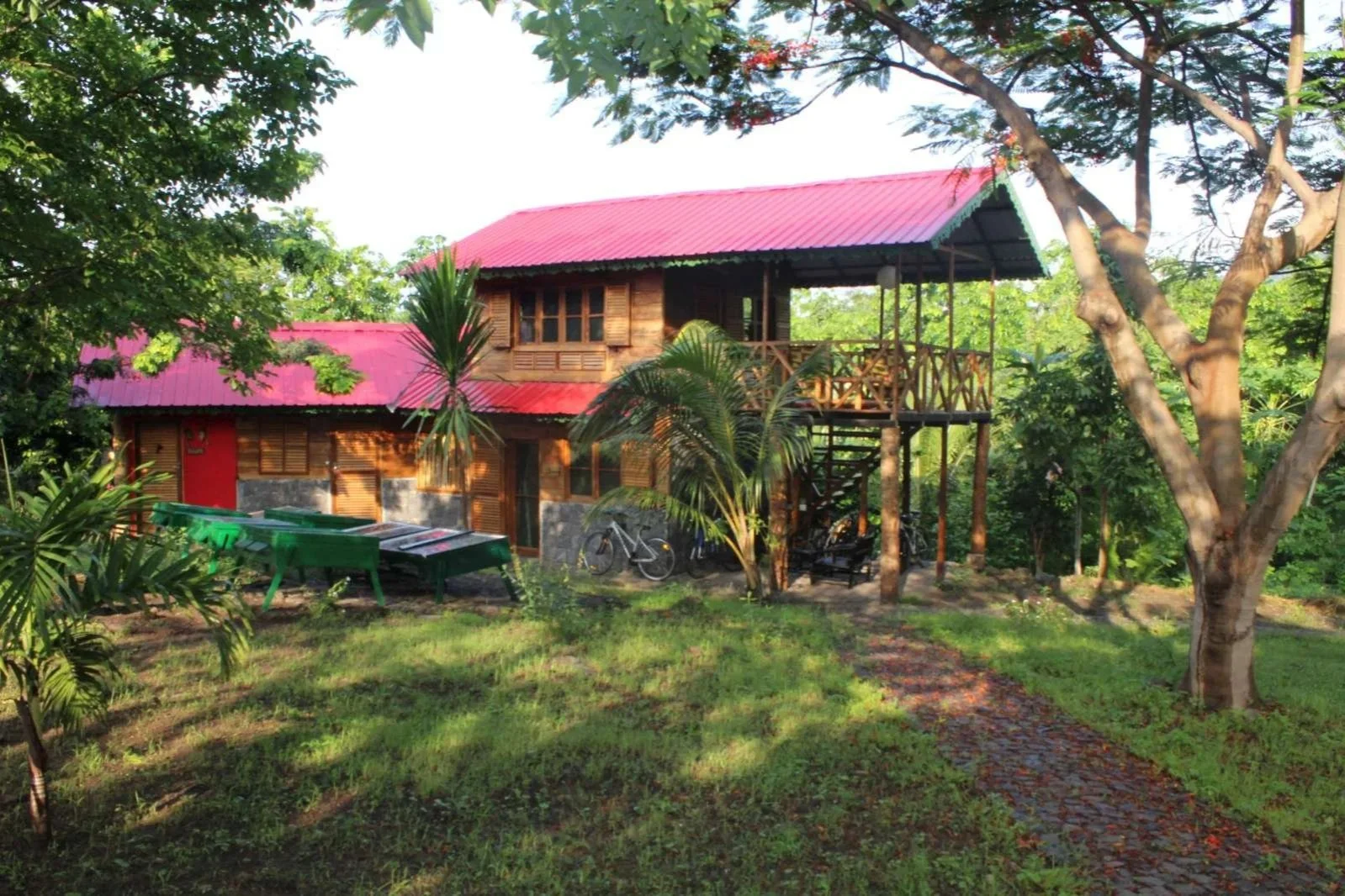Maison en bois avec un toit en tôle rouge, entourée de verdure et d'arbres, avec un vélo et des chaises en plastique devant.