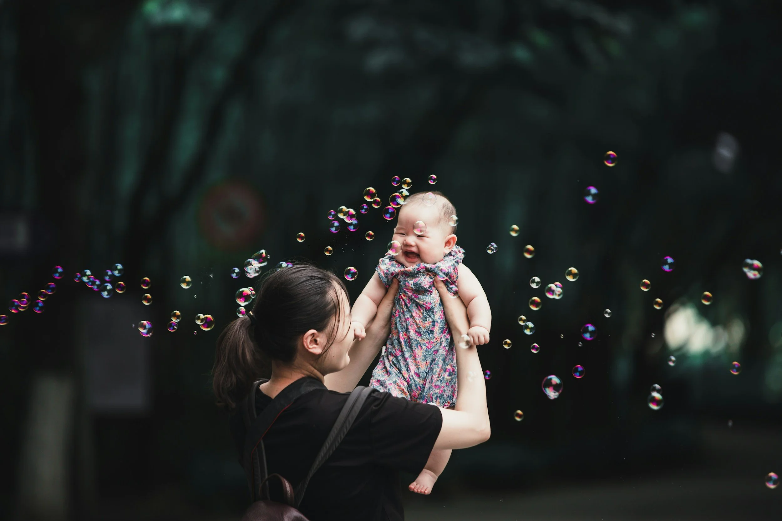A woman holding a smiling baby girl high in the air among floating soap bubbles outdoors surrounded by dark trees.