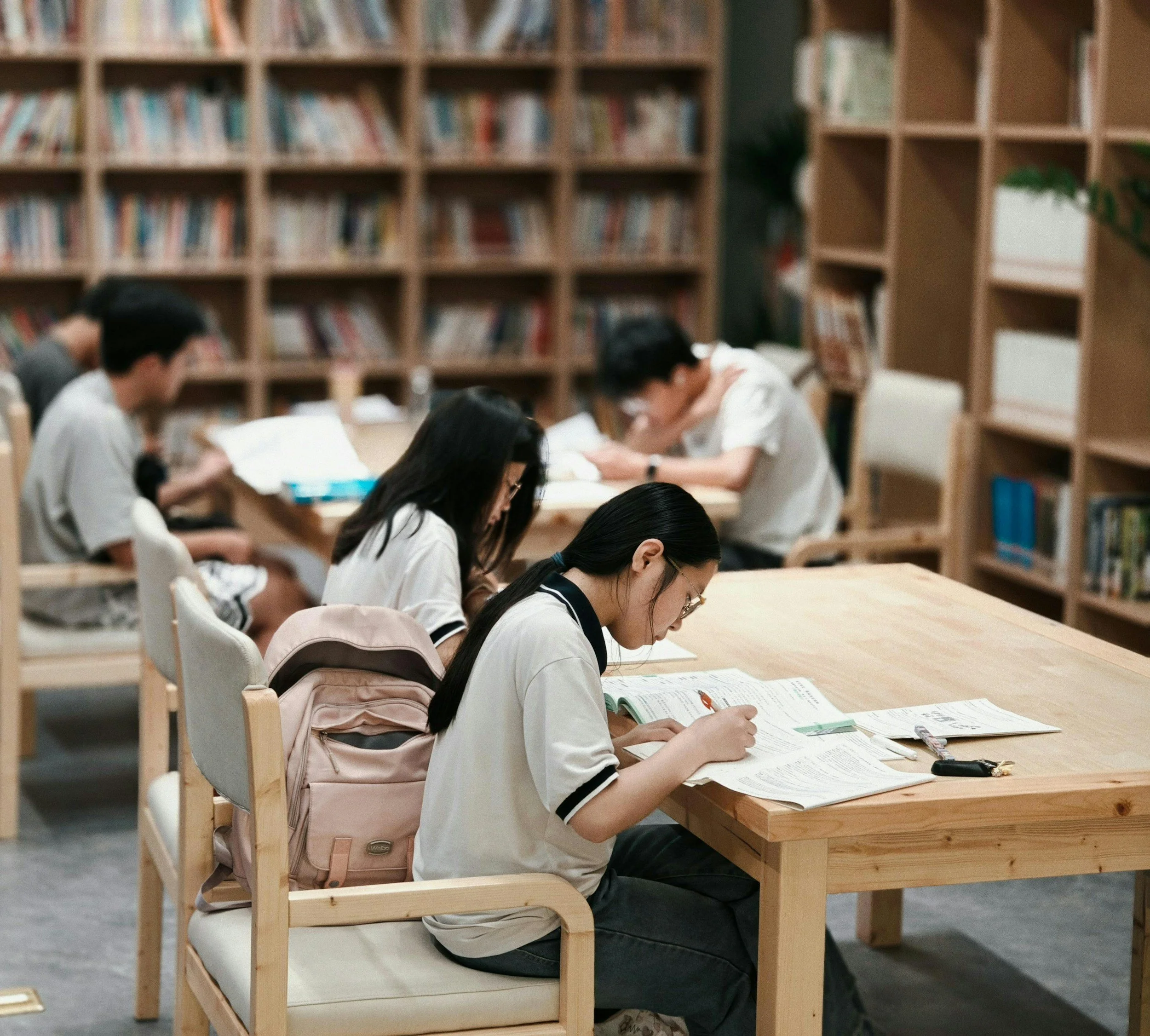 Young students sitting at a wooden table reading books in a library with wooden bookshelves in the background.