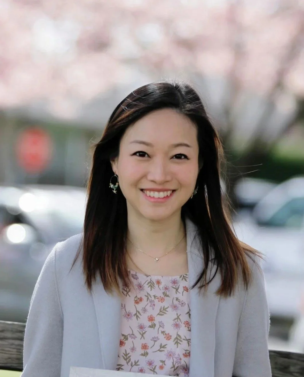 A woman with long dark hair smiling outdoors with cherry blossoms in bloom in the background.