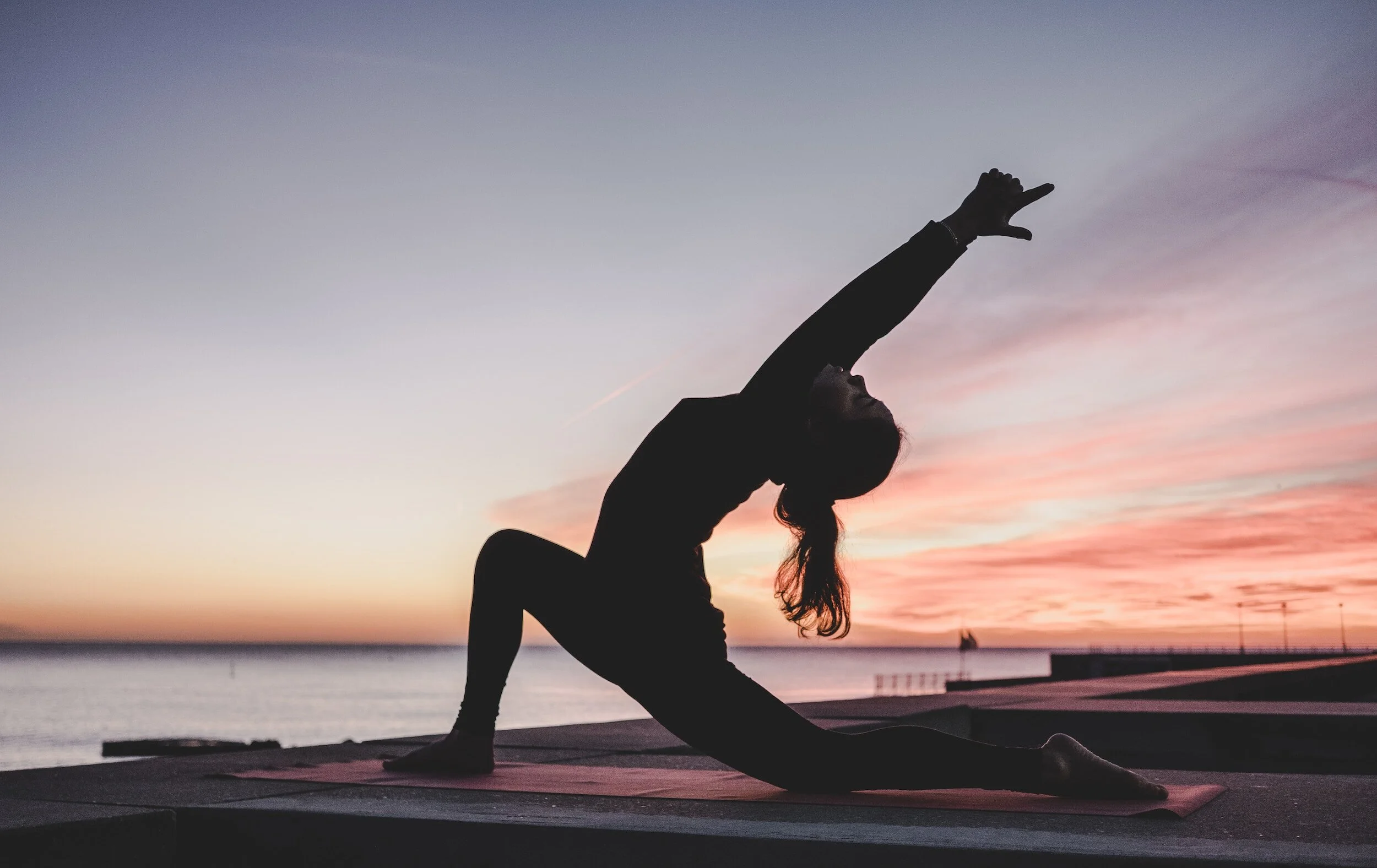 Silhouette of a person practicing yoga on a mat at sunset by a body of water, with a colorful sky in the background.
