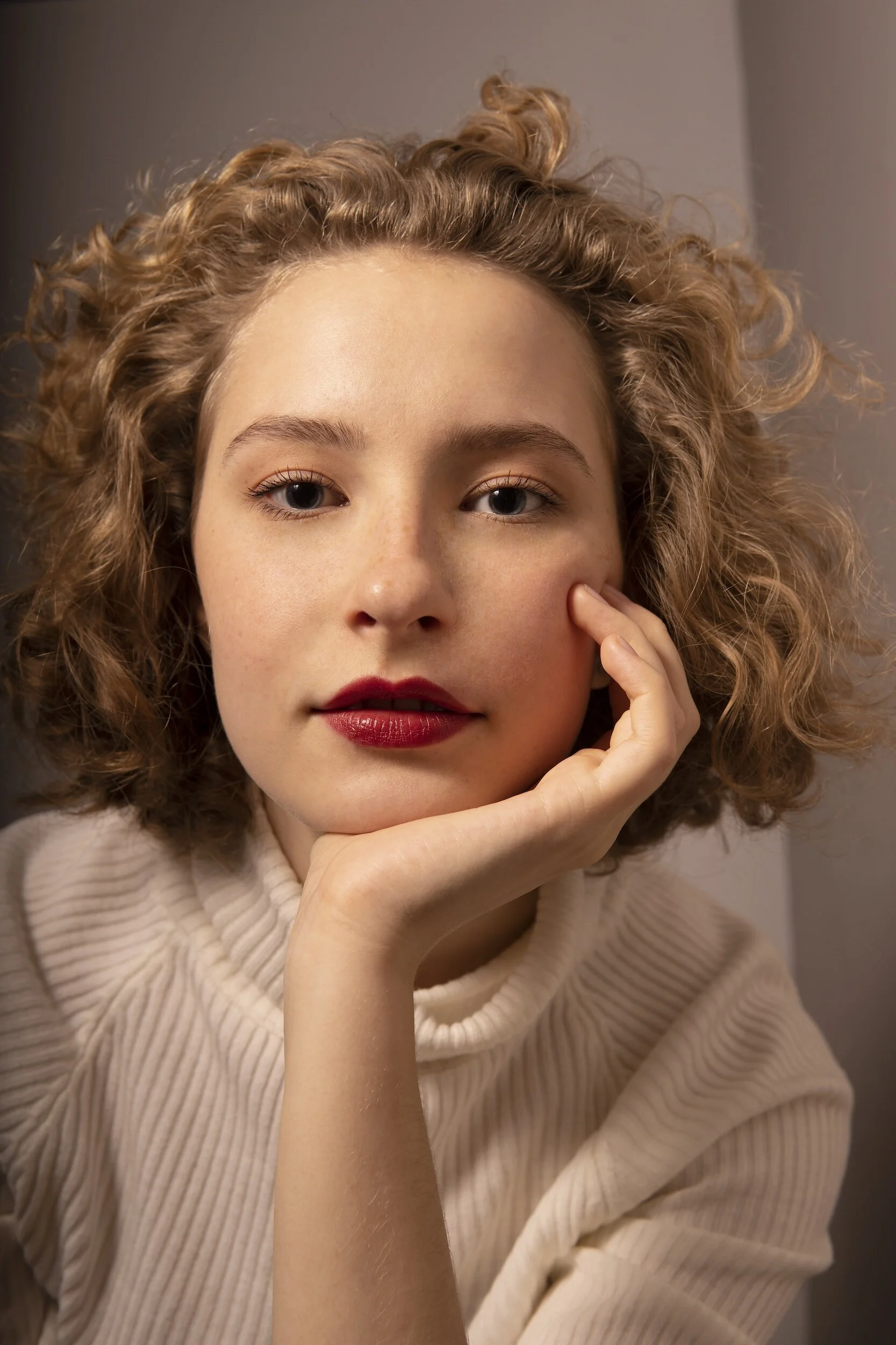 Woman with curly hair and red lipstick, wearing a cream turtleneck, posing with hand on cheek.