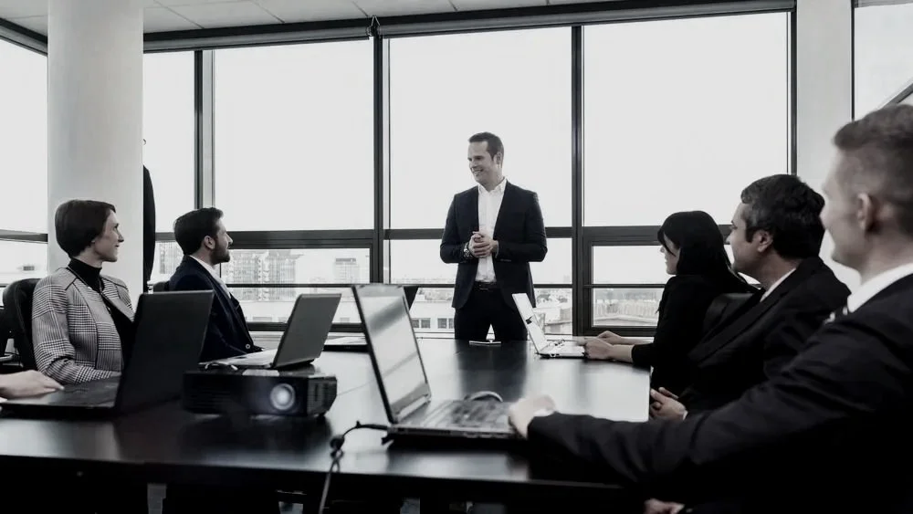 A well-dressed man in a suit jacket is addressing a table of colleagues who have laptop computers in front of them. large, bright windows of a modern office form the backdrop.