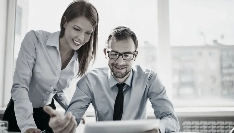 A professionally-dresed young woman stand next to a seated young man in glasses wearing a dress shirt and tie peering at an open laptop computer screen.