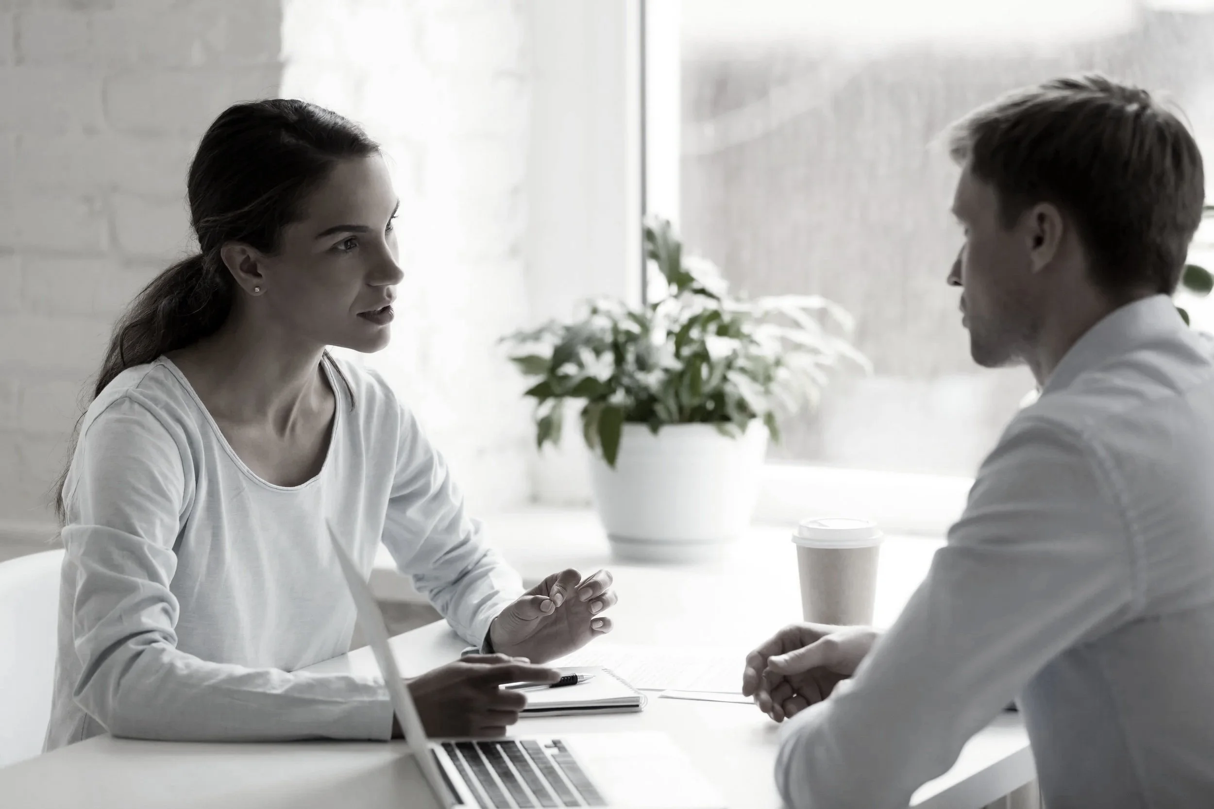A youg man and woman sit across from each other at a table in conversation. A cofe cup is near the man with some papers scattered on the table. A potted plant is in the background.