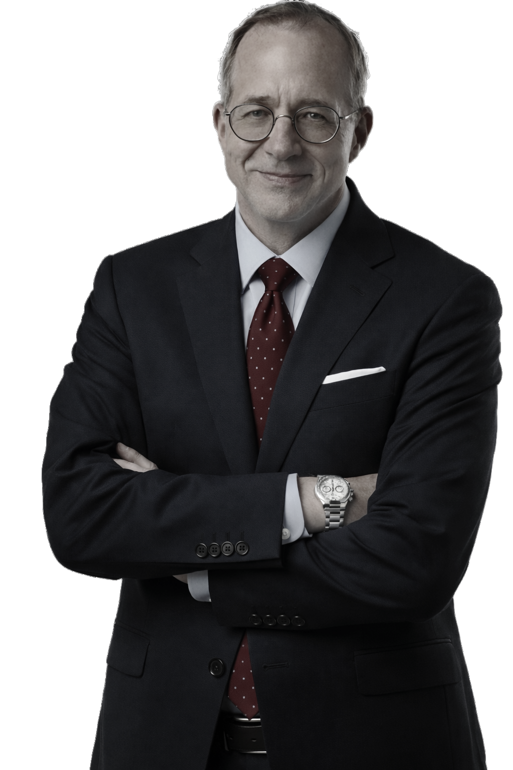 A well-dressed businessman with short hair and glasses is standing against a white background wearing a blue suit, light-colored shirt, and red tie with arms folded.