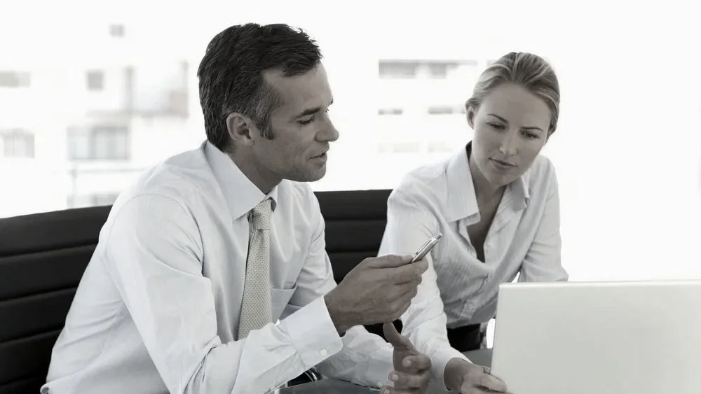 A well dresed man and woman are seated at a desk pering at an open laptop computer. The man is gesturing toward the computer screen with pen in hand.