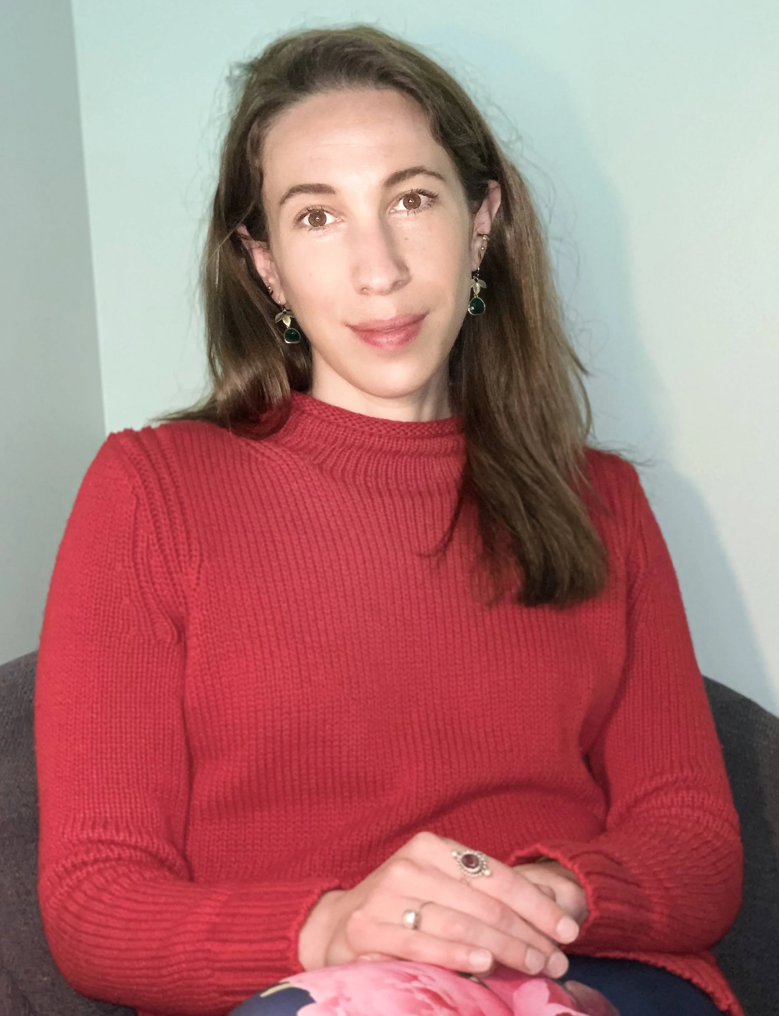 A woman with long brown hair wearing a red sweater and floral pants, sitting on a chair against a light green wall.