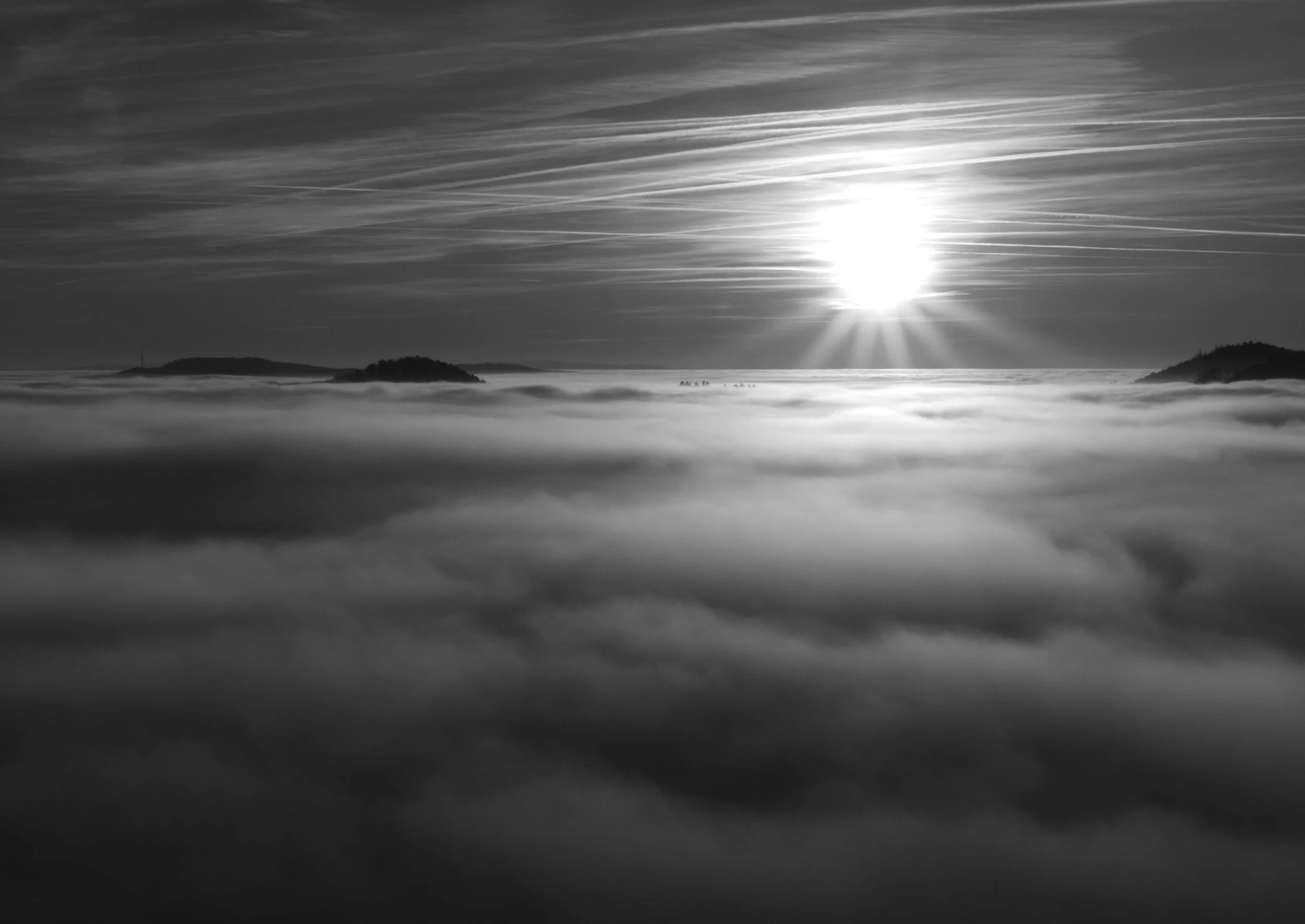 Black and white photo of a landscape with the sun shining over cloud-covered mountains and small islands in the distance.