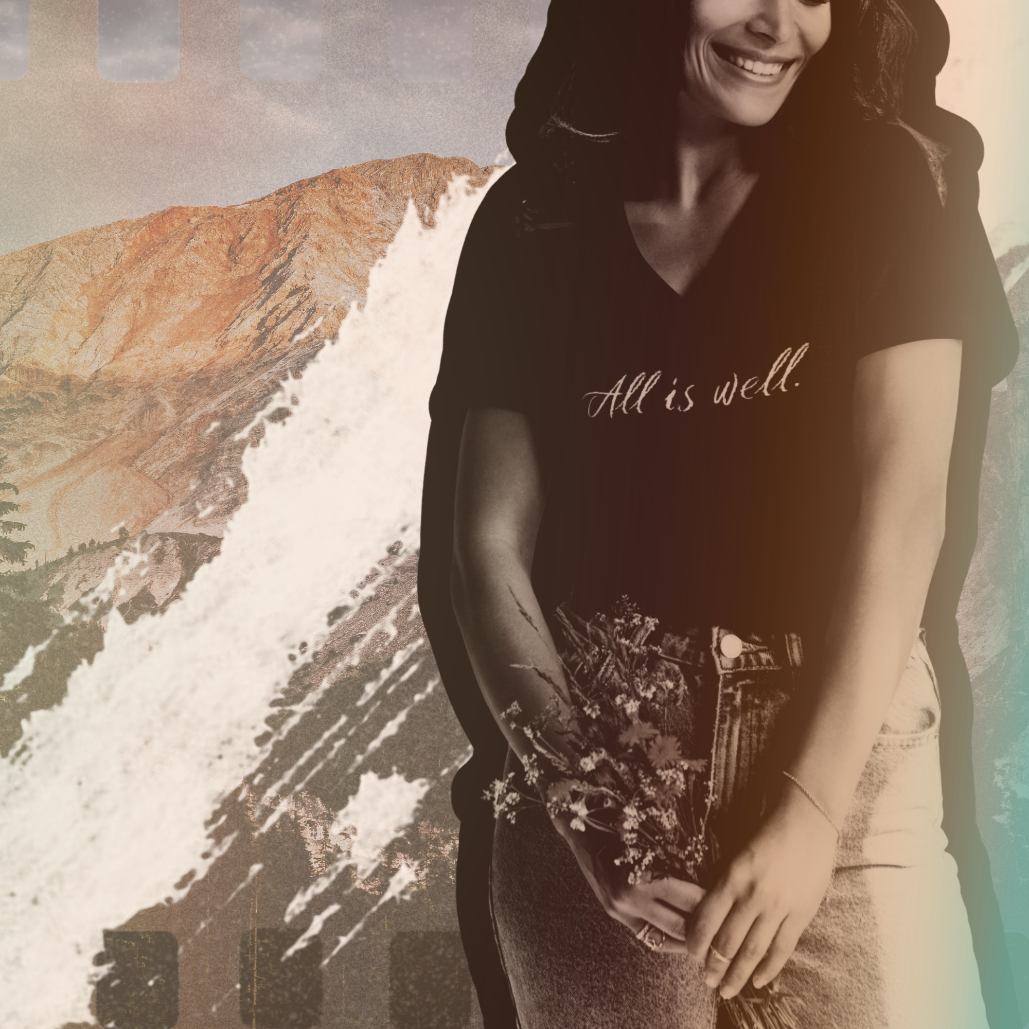 A smiling woman with dark hair is wearing a black t-shirt that says 'All is well.' and light-wash jeans with a floral pattern. She is standing in front of a mountainous background with a waterfall.