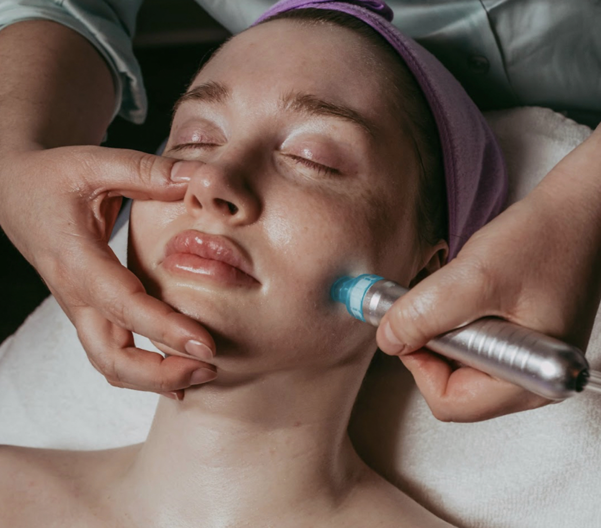 A woman with her eyes closed receiving a facial treatment with a cosmetic tool on her cheek from a professional.