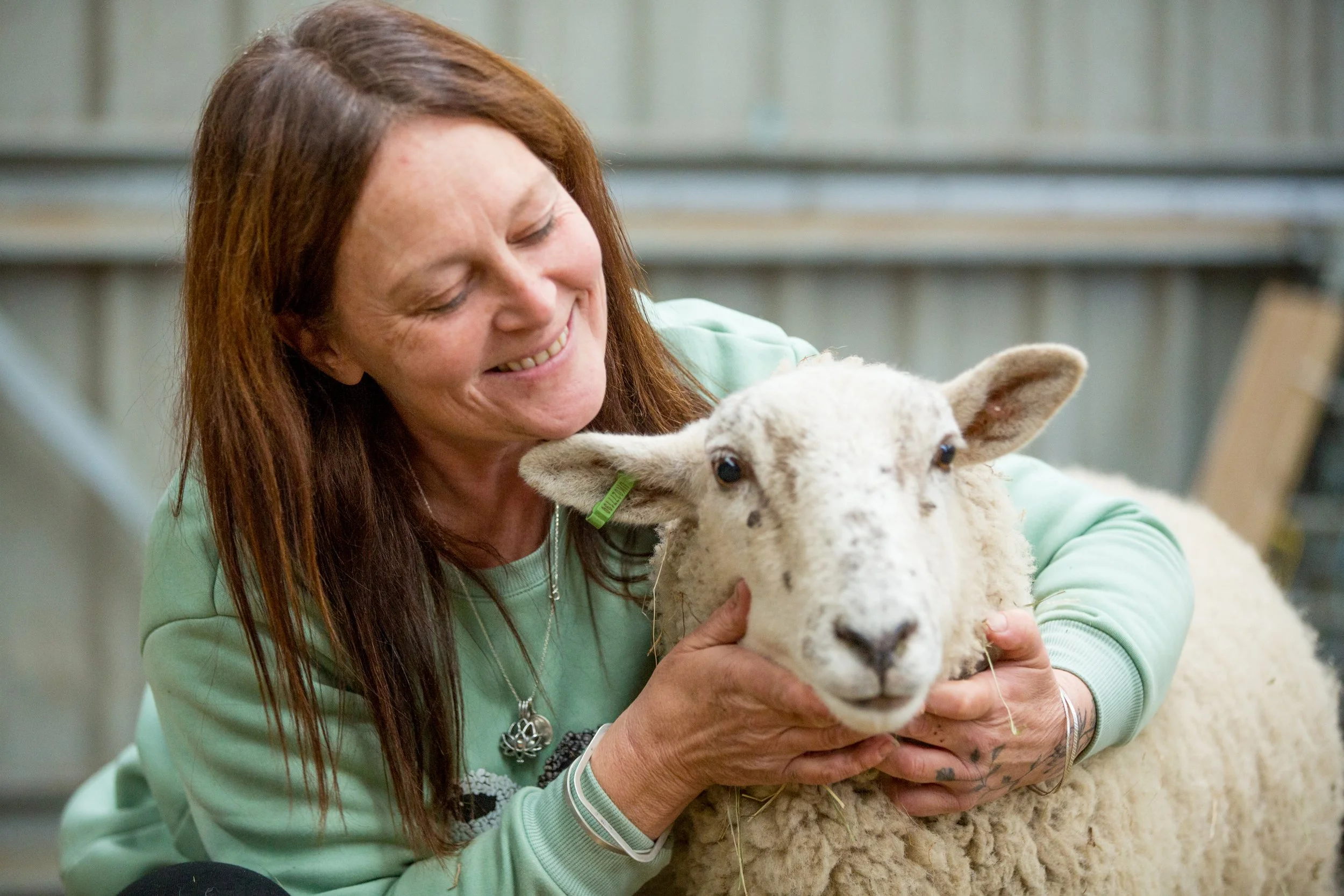 A woman with long reddish hair smiling while holding a young sheep in a barn or farm setting.