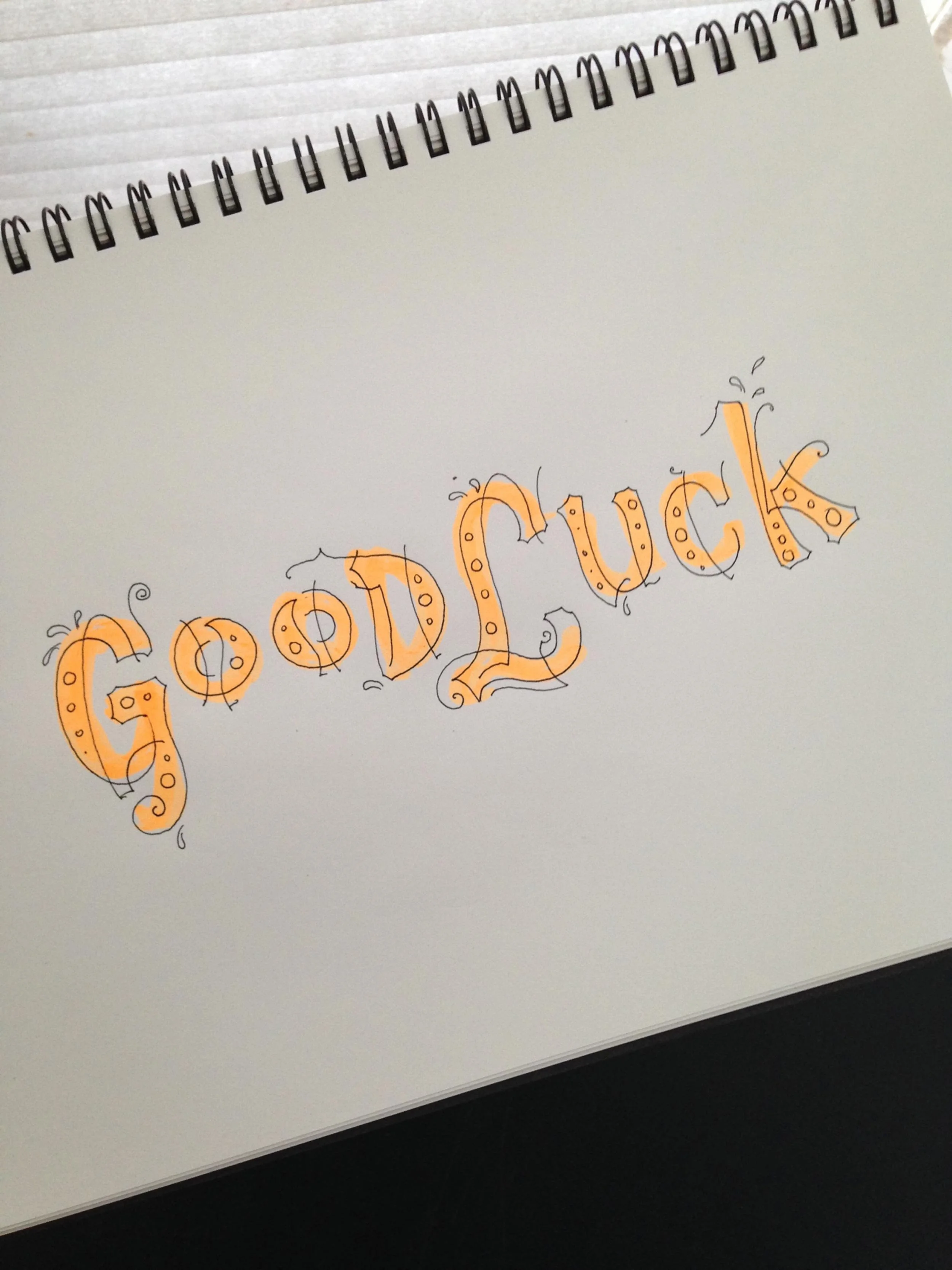 Sketchbook showing hand-drawn "Good Luck" in decorative orange letters with black outlines and swirls.