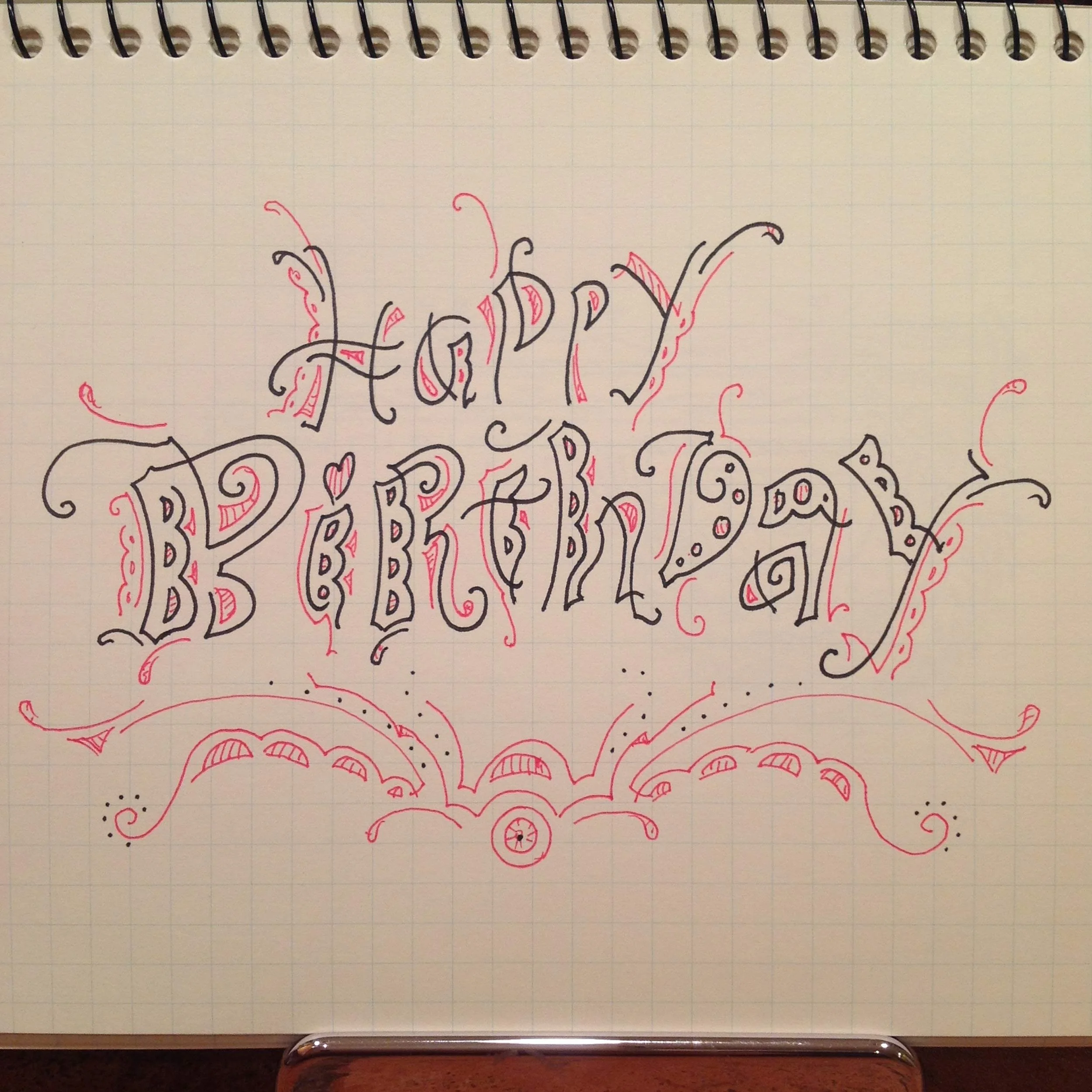 Stylized handwritten text on grid paper reading 'Happy Birthday' with decorative red and black designs.