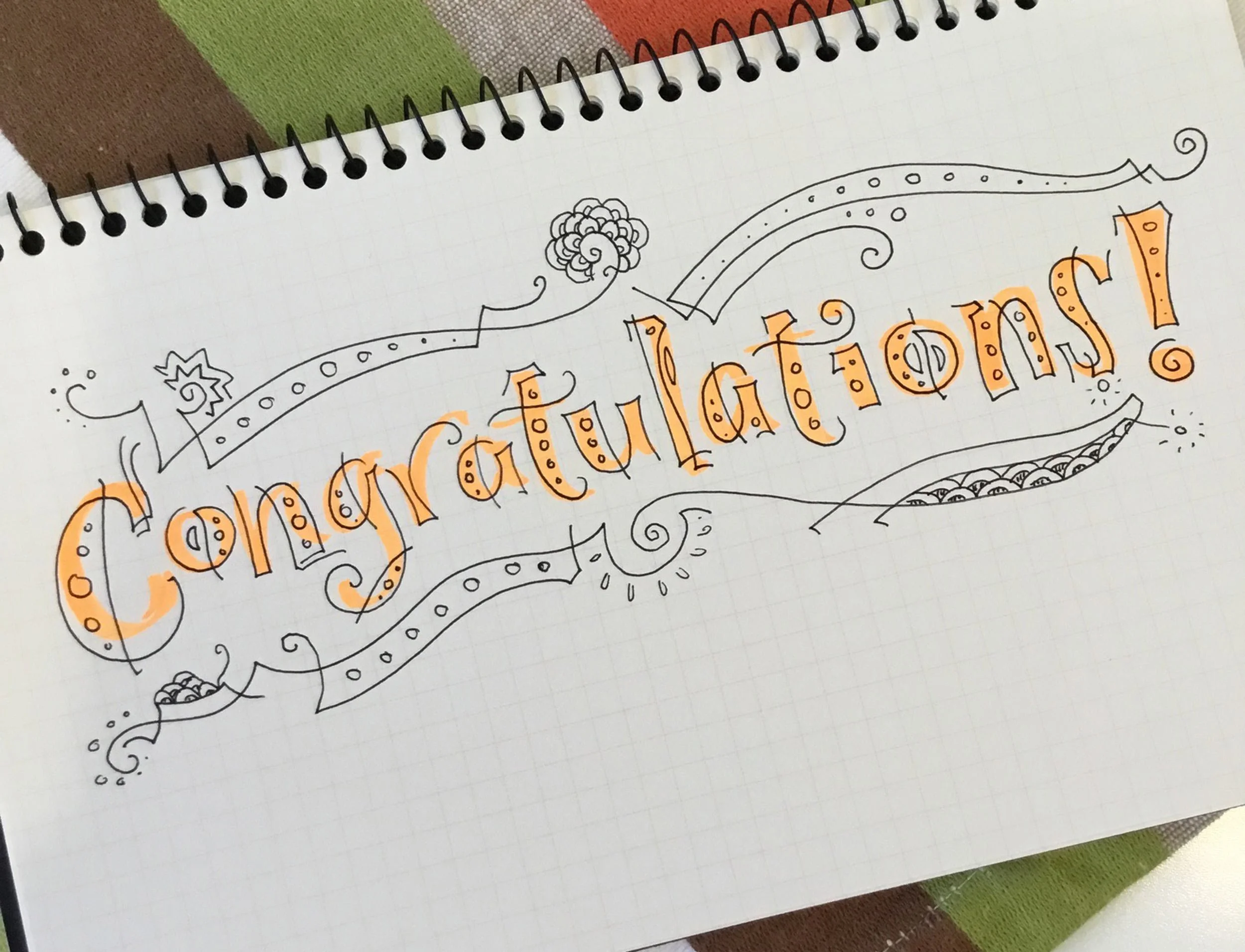 Doodle artwork spelling 'Congratulations!' in orange and black with decorative elements on a notebook.
